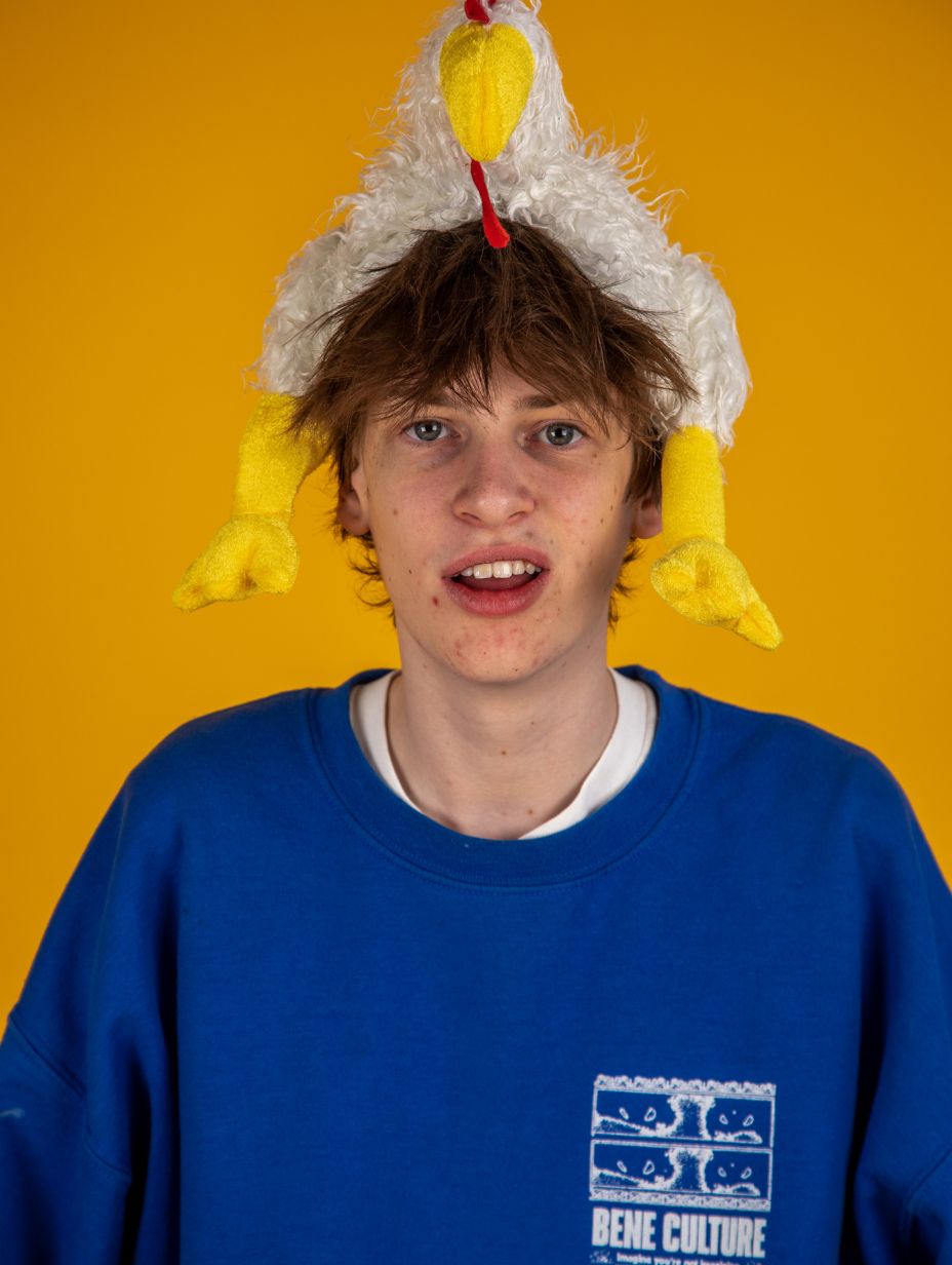 London comedian Jon Gibbons in a promotional headshot, wearing a novelty stuffed chicken hat and a bright blue sweatshirt, looking directly at camera against a vivid yellow studio backdrop with an open expression.