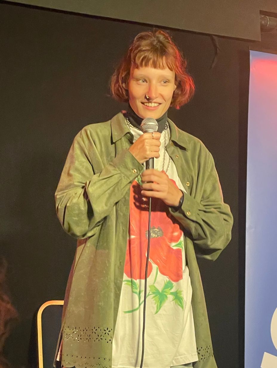 London comedian Verity Sharpe holds a microphone in both hands and smiles at a comedy show, wearing an oversized olive jacket over a white poppy print t-shirt, short reddish hair, warm red stage light behind them. 242 characters ✓