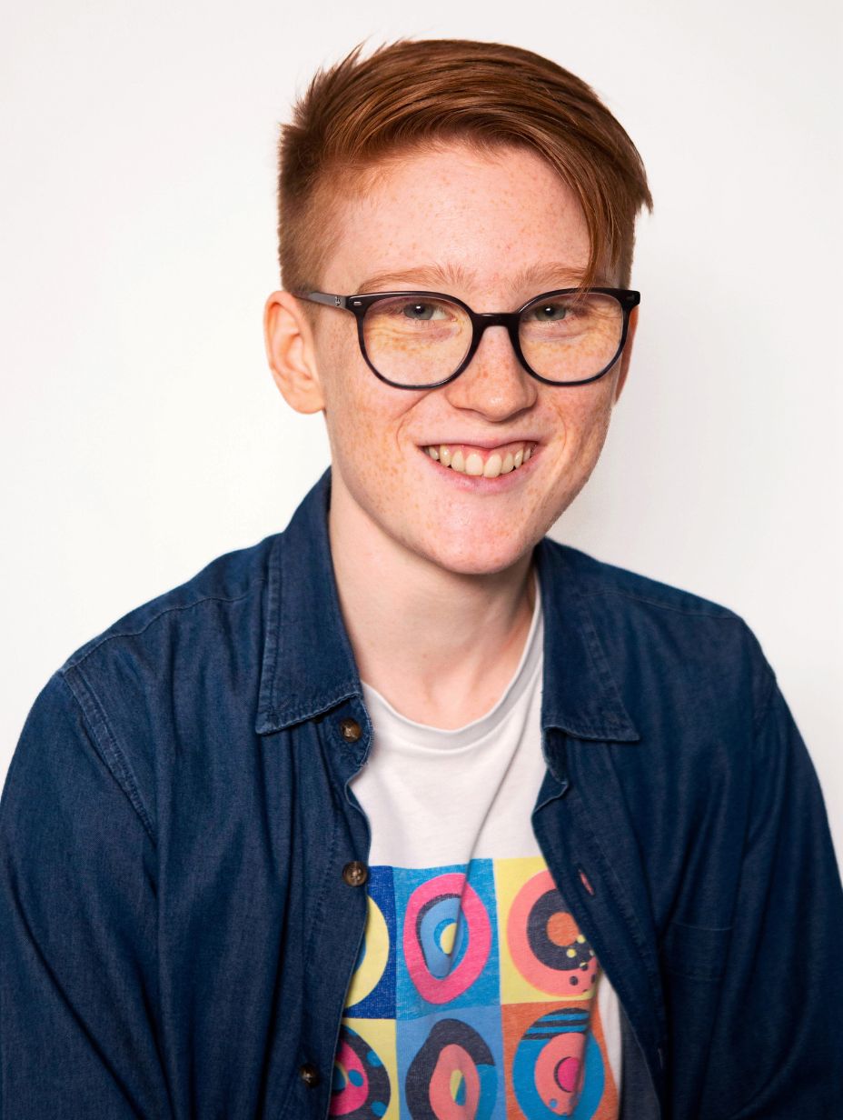 London comedian Ro Dodgson smiling in a studio headshot, wearing a blue denim shirt over a colourful graphic tee, red hair swept to the side, dark frame glasses, and a warm open smile against a clean white backdrop.