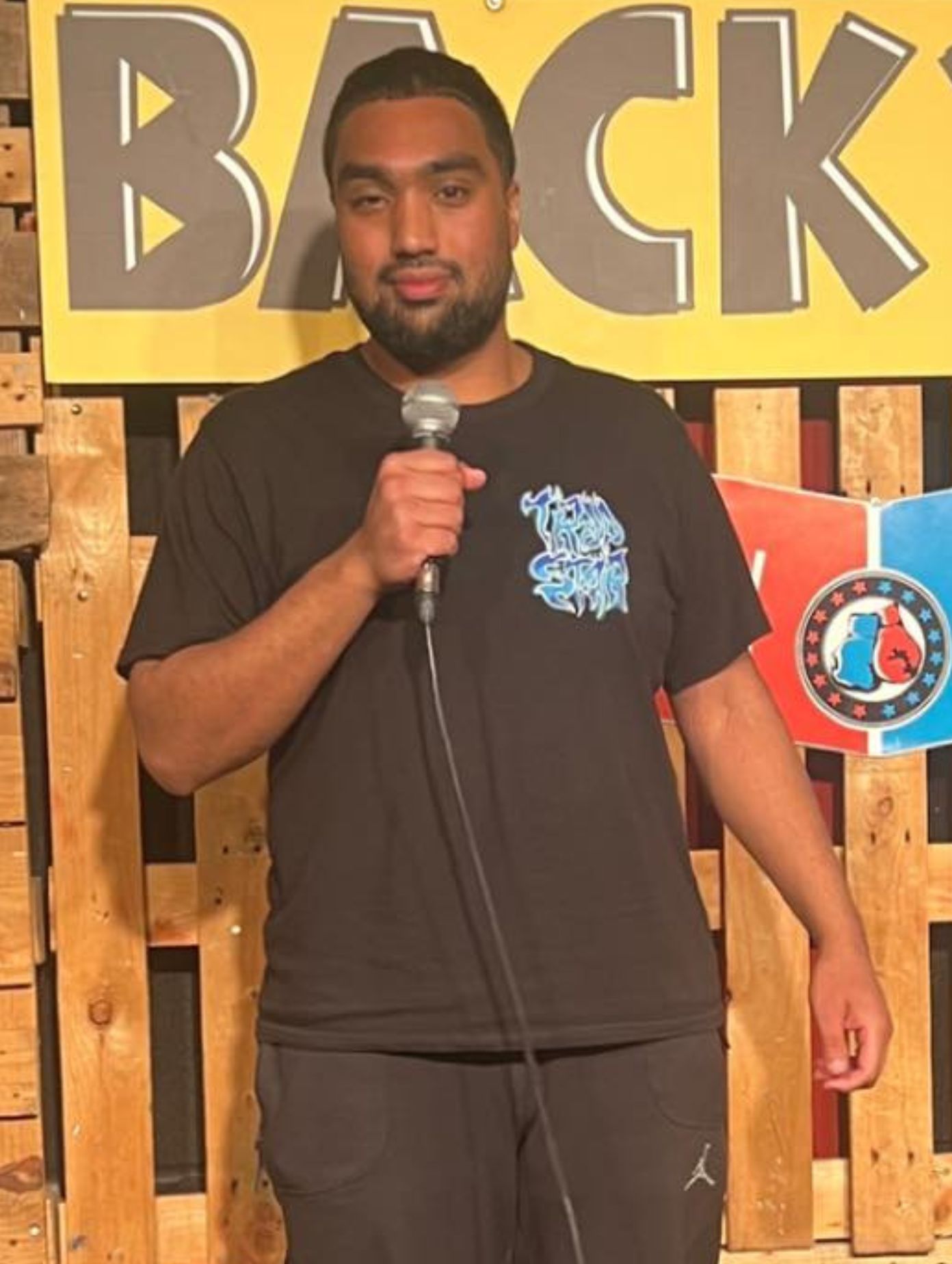 London comedian Abdul Mustakim on stage in a black graphic T-shirt and black joggers, holding a silver microphone at a comedy club, wooden backdrop and bold competition signage visible behind him under stage lighting.