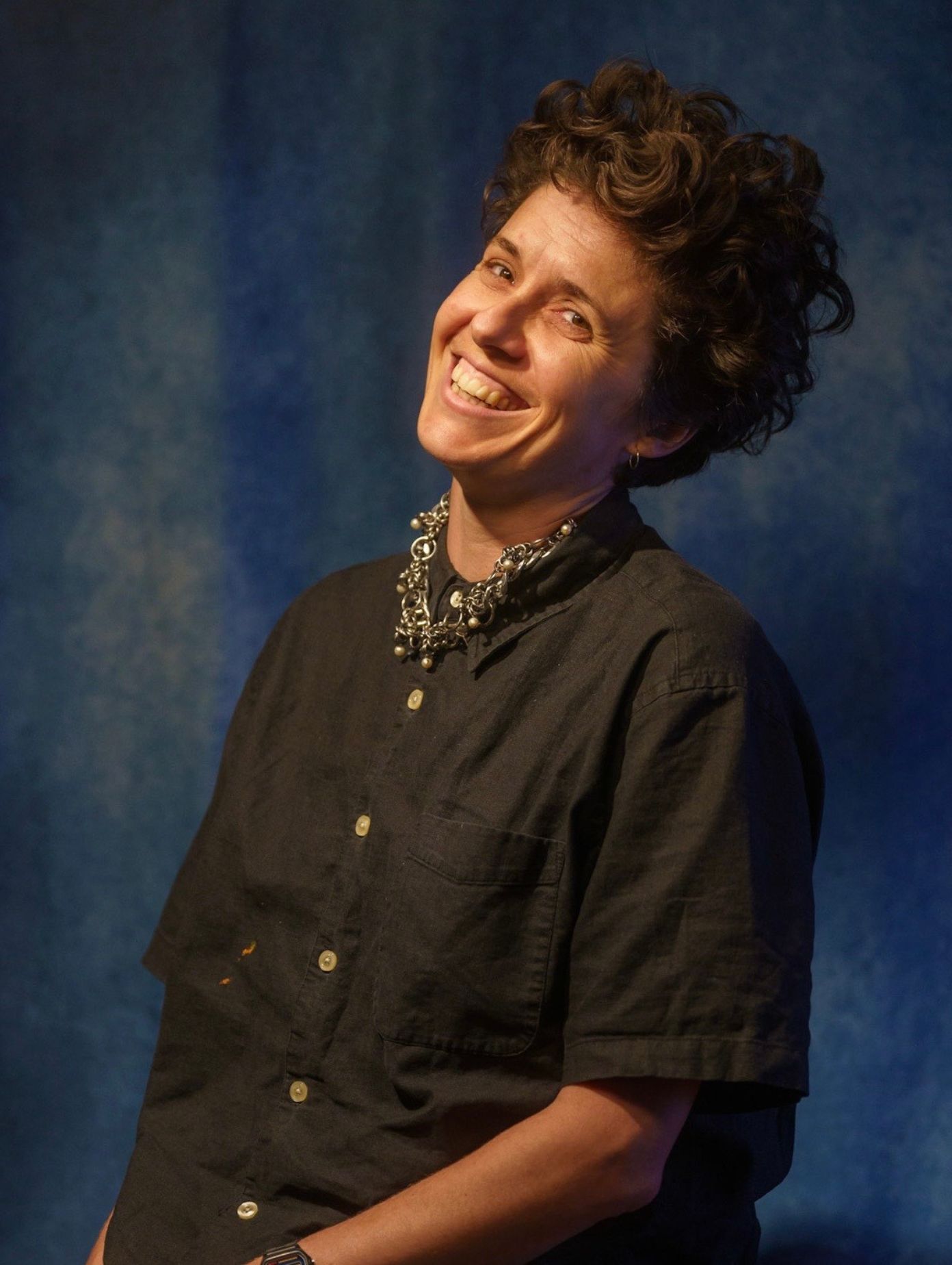 London comedian Celine Kuklowsky laughing broadly in a promotional headshot, wearing a black linen shirt and silver chain necklace with short dark curly hair, photographed against a deep blue textured backdrop.