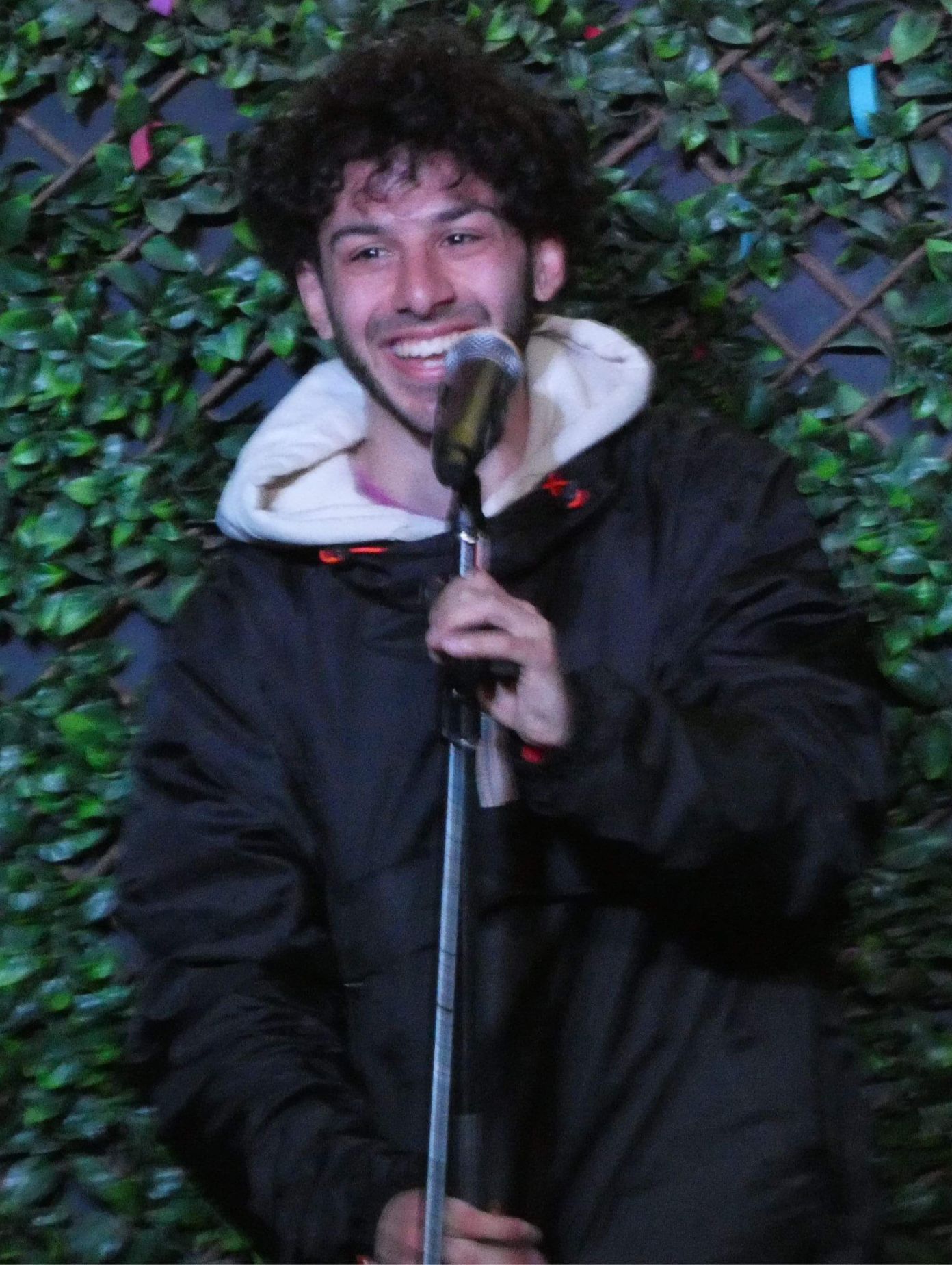 British-Iraqi comedian Sammer Elzubaidi beaming on stage at a comedy club in London, holding a vintage microphone stand in a black jacket over a white hoodie, a decorative green ivy trellis wall with colourful confetti pieces visible behind him.