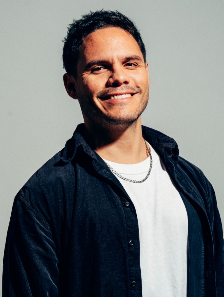 London comedian Héctor Ayala smiling in a professional headshot, wearing a dark navy shirt over a white tee and silver chain necklace, photographed against a clean light grey background with warm natural lighting.