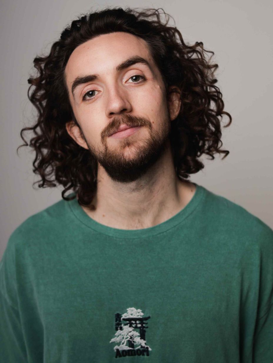 London comedian Darren Cullen in a studio headshot, wearing a green Aomori T-shirt with long dark curly hair and light stubble, looking up with a knowing expression against a clean neutral grey background.