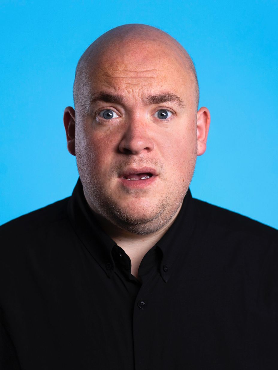 London comedian Jim Hooker in a promotional studio headshot, bald-headed with wide eyes and mouth slightly open in a comedic expression, wearing a black button-down collar shirt against a vivid cyan blue backdrop.