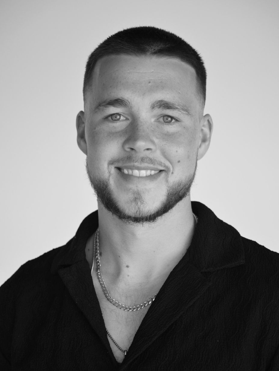London comedian Finn Feeney in a black and white studio headshot, wearing a black open-collar shirt and silver chain necklace, short faded hair and light stubble, smiling warmly against a clean white background.