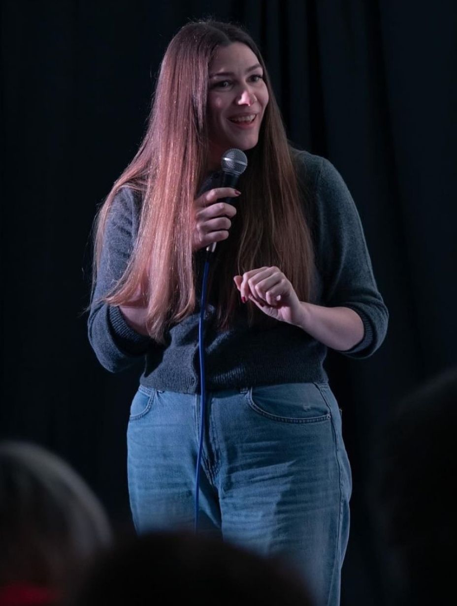 London comedian Claire Cox mid-set at a London comedy club, holding a microphone in a dark grey knit sweater and light blue jeans, long straight hair with pink highlights catching the stage lighting against a dark black curtain backdrop.