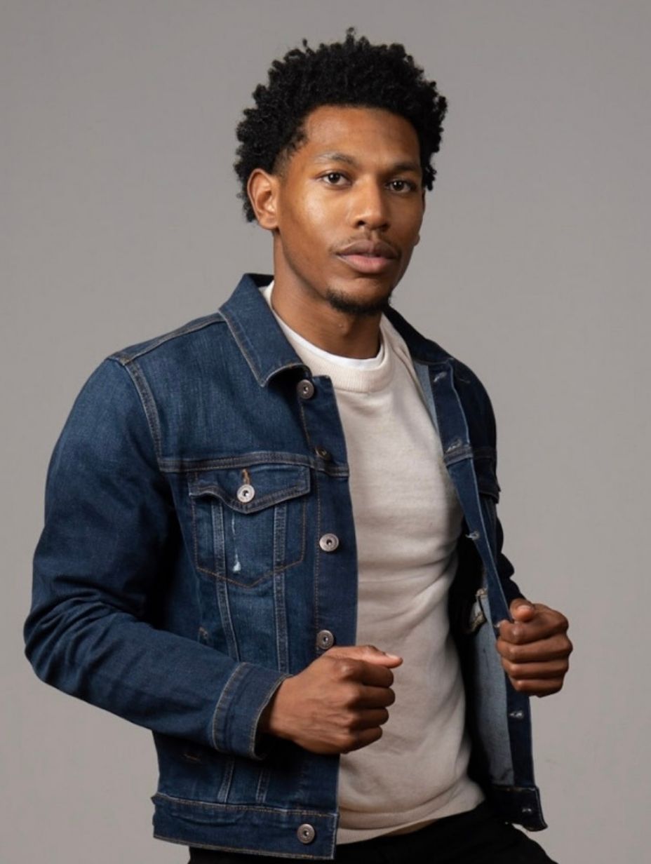London comedian Derrick Carter in a promotional headshot, wearing a blue denim jacket open over a white knit sweater, short natural hair, holding the lapels of his jacket against a neutral grey studio background.