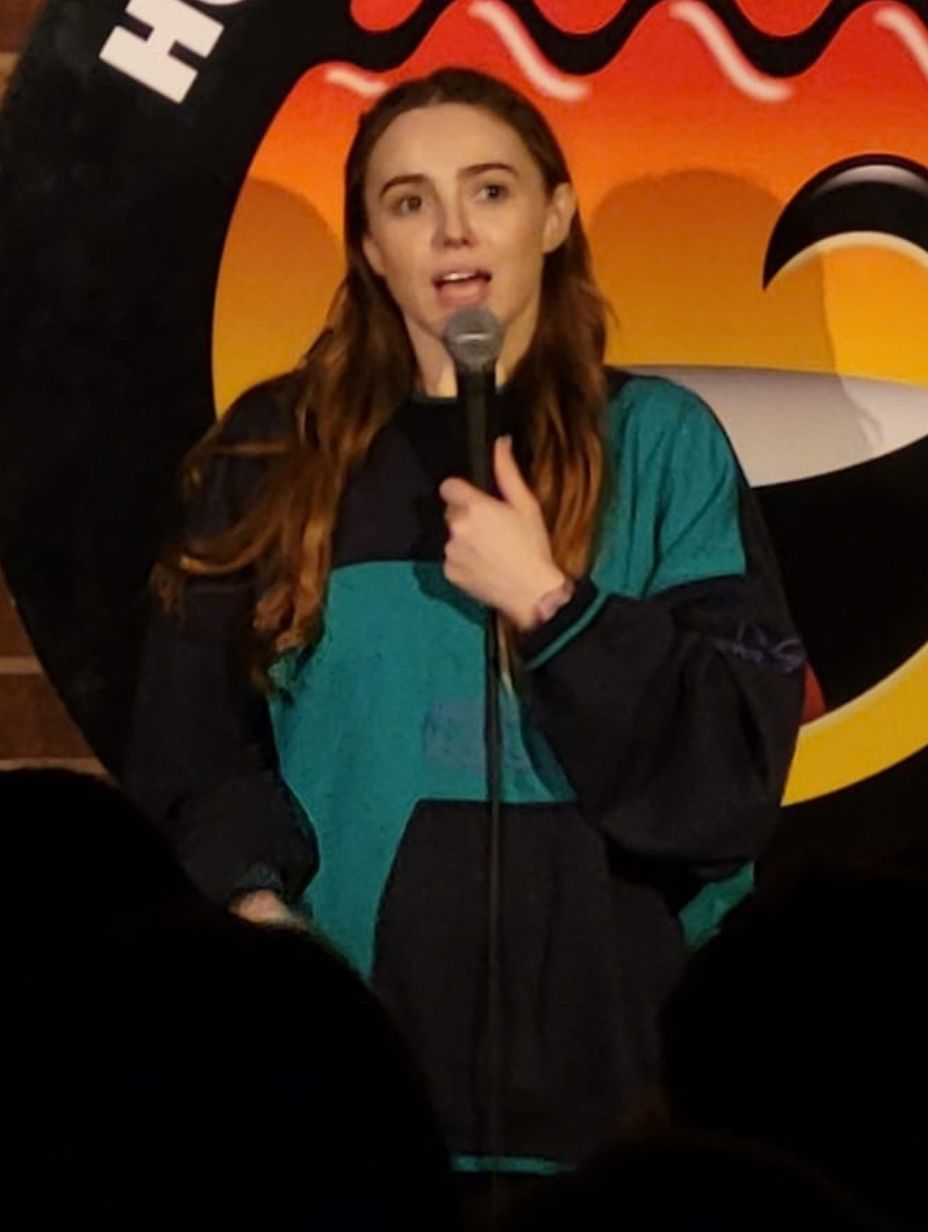 London comedian Kat Ronson performing on stage, holding a microphone in a black and teal vintage track jacket, long auburn hair and mid-set expression, large circular comedy logo visible on brick wall behind her.