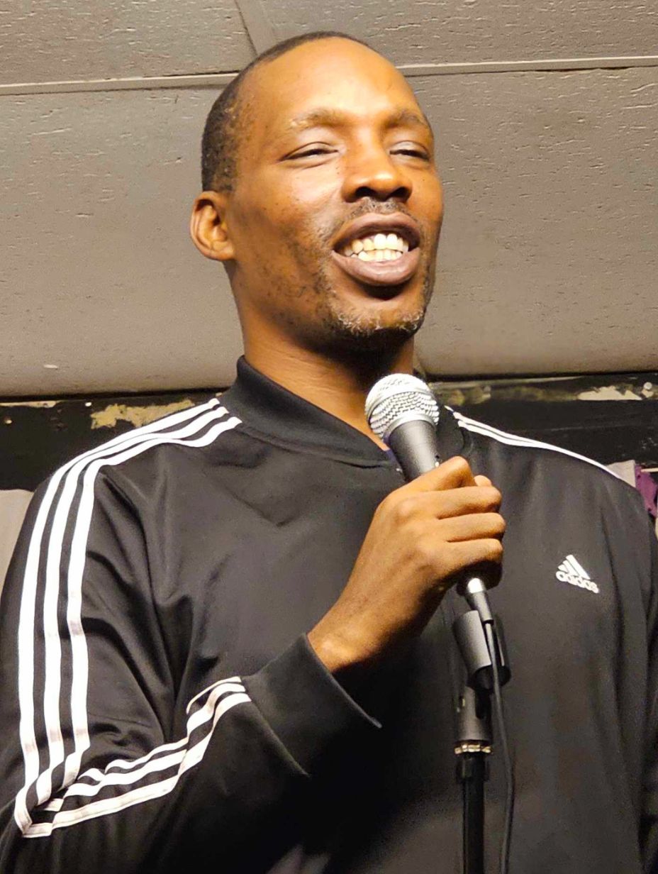 London comedian Emmanuel Paul performing on stage, wearing a black Adidas sports jacket while holding a microphone, head tilted back with an open joyful expression. 