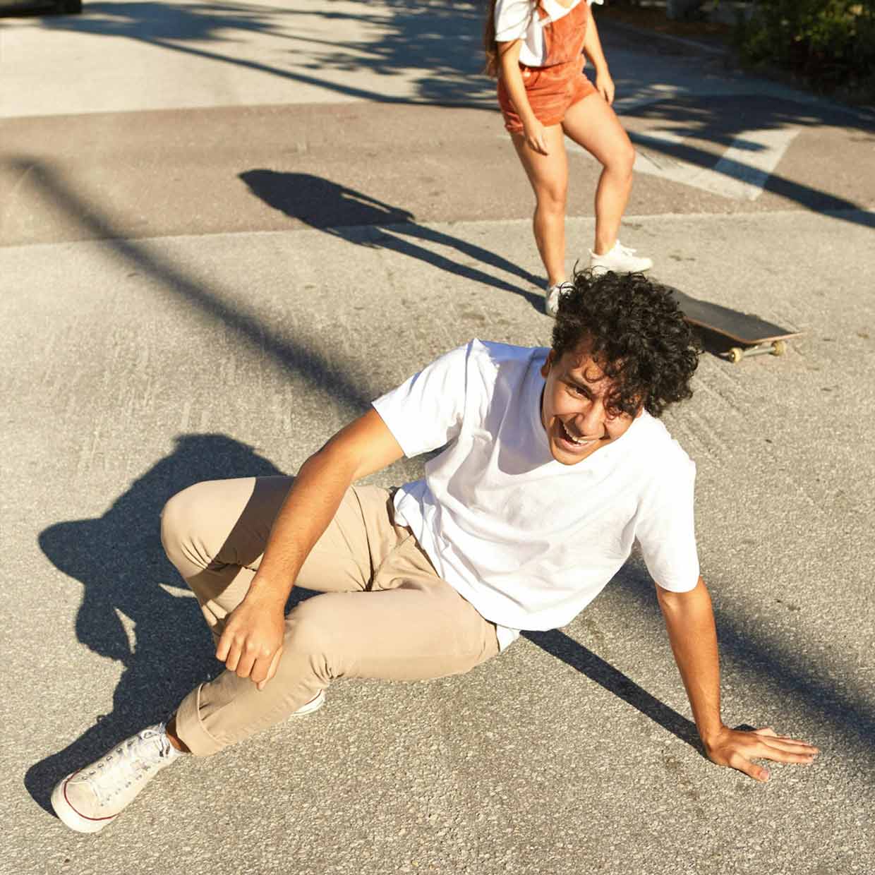 Ein junger Mann sitzt lachend auf der Straße, neben ihm ein Skateboard. Im Hintergrund steht eine junge Frau. Die Szene zeigt Leichtigkeit, Freundschaft und den Spaß an Bewegung an einem sonnigen Tag.