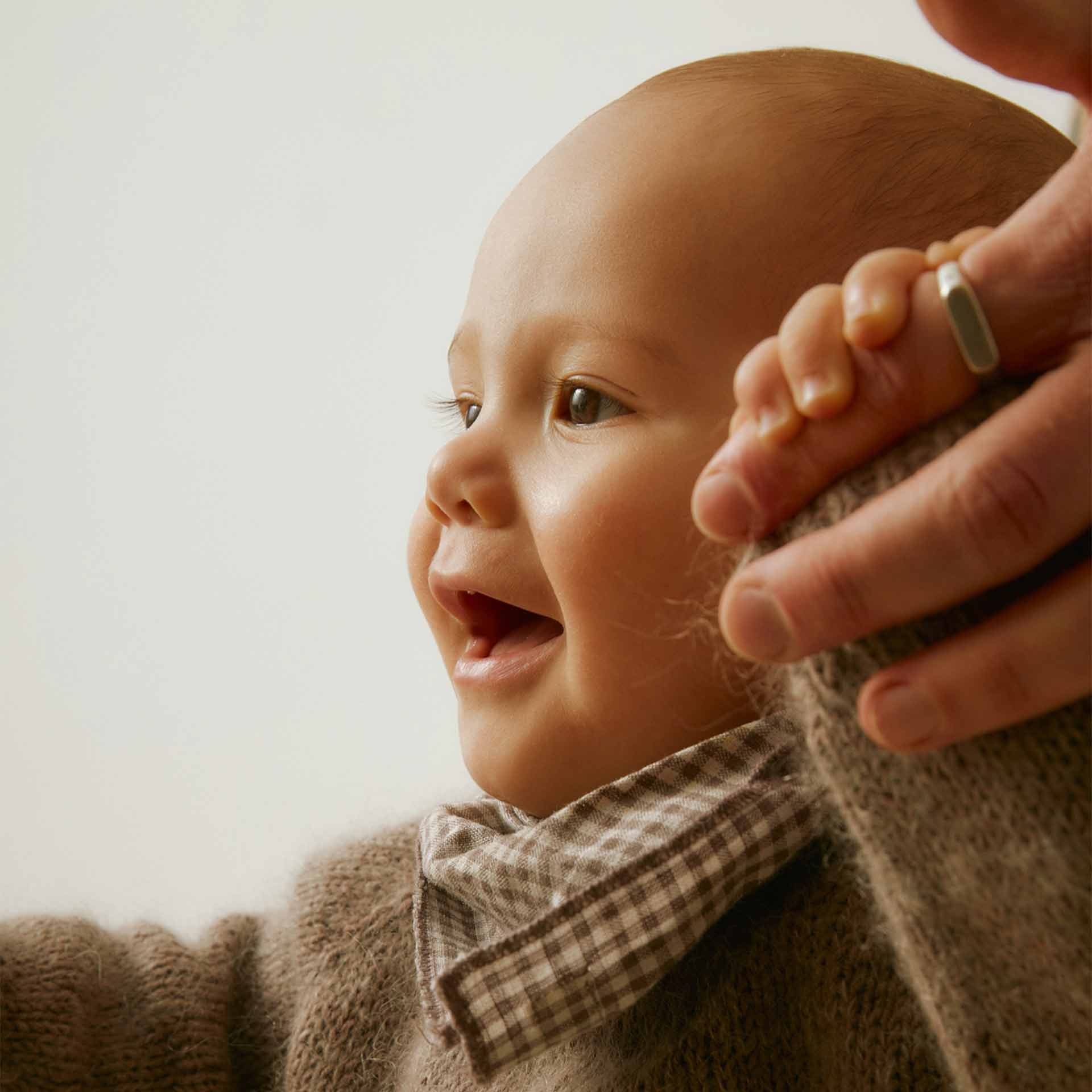 Ein fröhliches Baby lächelt strahlend und hält liebevoll die Hand eines Erwachsenen. In weicher Kleidung gekleidet, vermittelt das Bild Wärme, Geborgenheit und die Magie früher Kindheitsmomente.