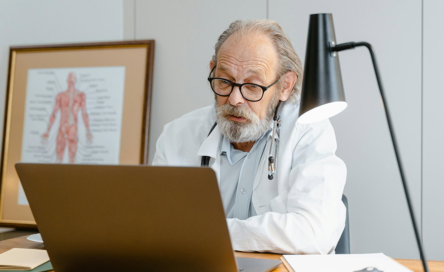 Ein älterer Arzt mit Brille und Stethoskop sitzt konzentriert vor einem Laptop. In einem modernen Praxisraum führt er offenbar eine digitale Sprechstunde durch – Erfahrung trifft auf Telemedizin.