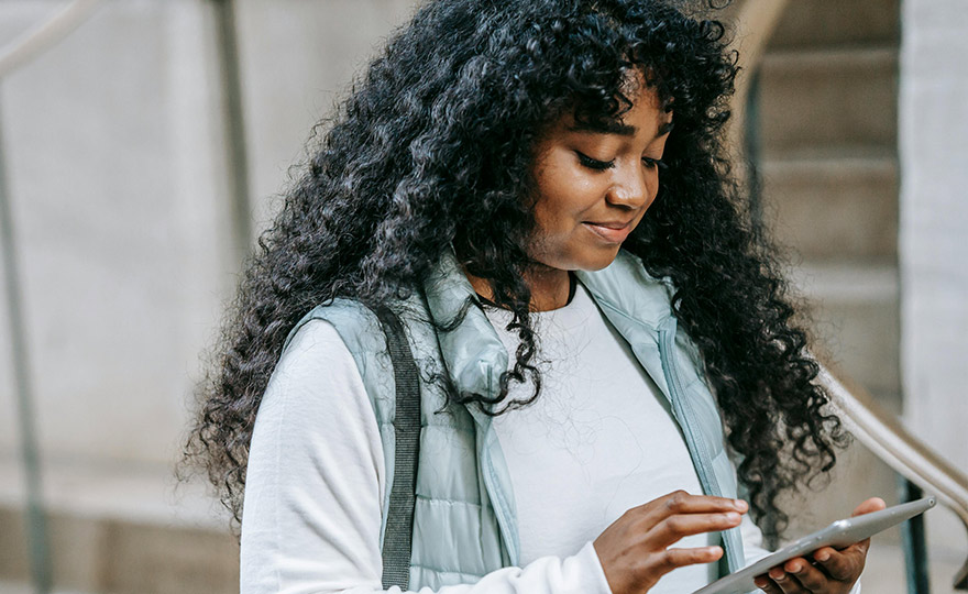 Eine junge Frau mit lockigem Haar nutzt konzentriert ein Tablet im Freien. Mit einem leichten Lächeln und entspanntem Blick zeigt sie digitale Selbstbestimmung – verbunden, modern und unterwegs aktiv.