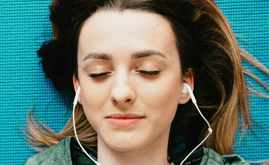 Eine junge Frau liegt entspannt mit geschlossenen Augen auf einer blauen Matte und hört Musik über Kopfhörer. Der Moment strahlt Ruhe, Achtsamkeit und innere Balance aus – ein Augenblick ganz bei sich.