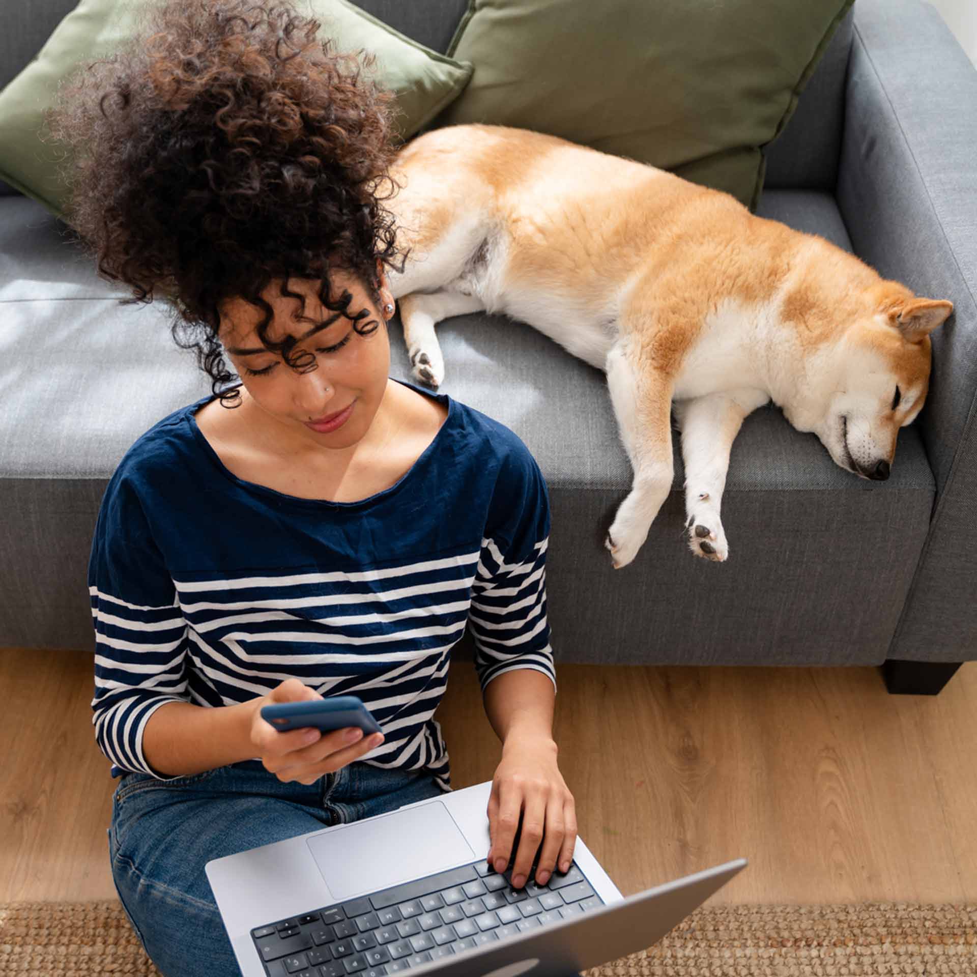 Frau arbeitet entspannt mit Laptop und Smartphone am Boden, während ihr Hund gemütlich auf dem Sofa schläft