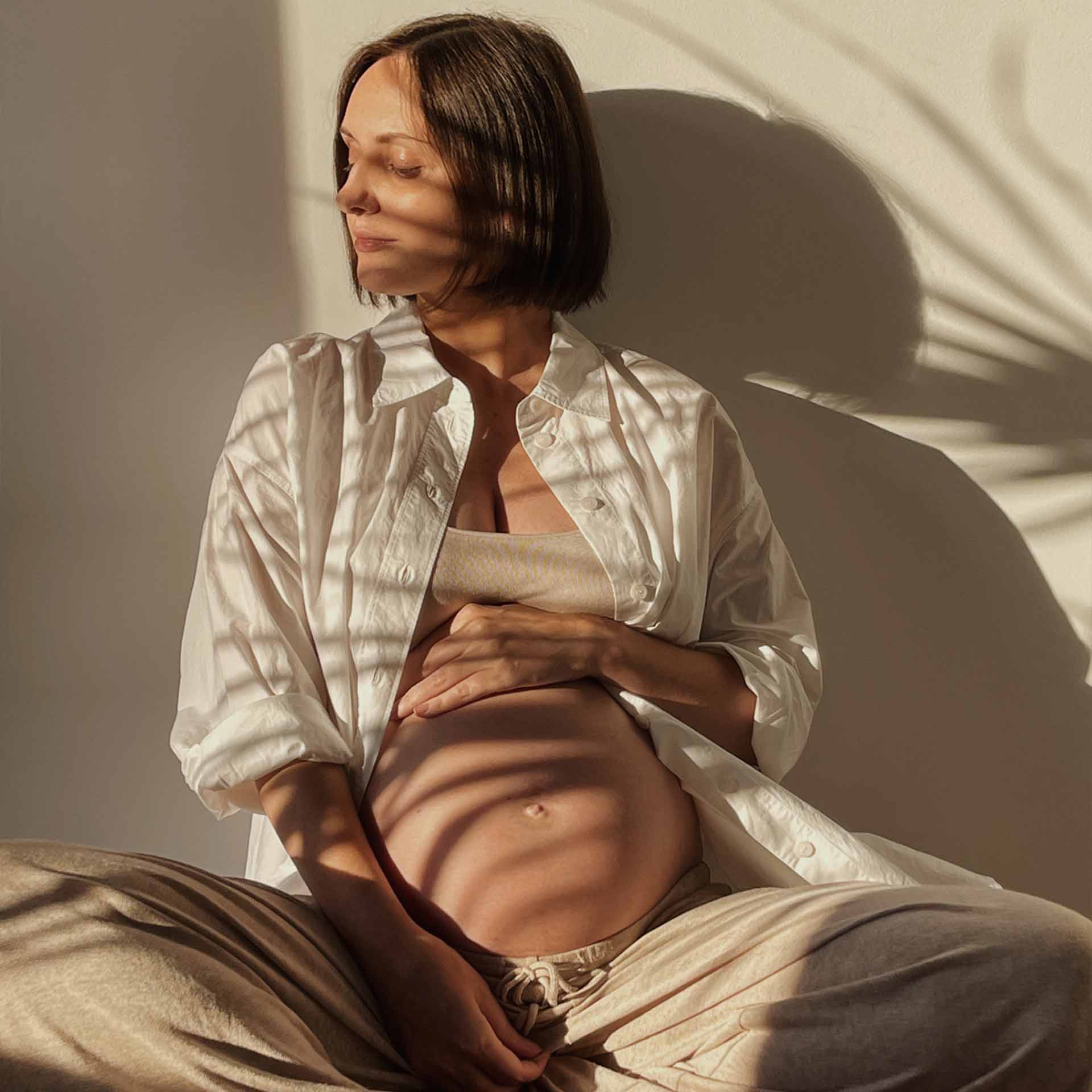 Eine schwangere Frau sitzt entspannt im Sonnenlicht und hält sanft ihren Babybauch. Sie trägt eine offene weiße Bluse, ein Top und bequeme Jogginghose. Weiche Licht- und Schattenspiele von Pflanzen spiegeln sich auf ihrem Körper und der Wand, was dem Bild eine ruhige und natürliche Atmosphäre verleiht.