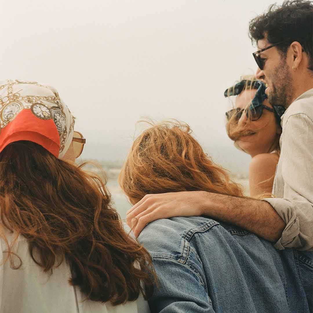 Vier Freunde stehen eng beieinander, schauen aufs Meer und genießen den Moment im warmen, windigen Wetter.