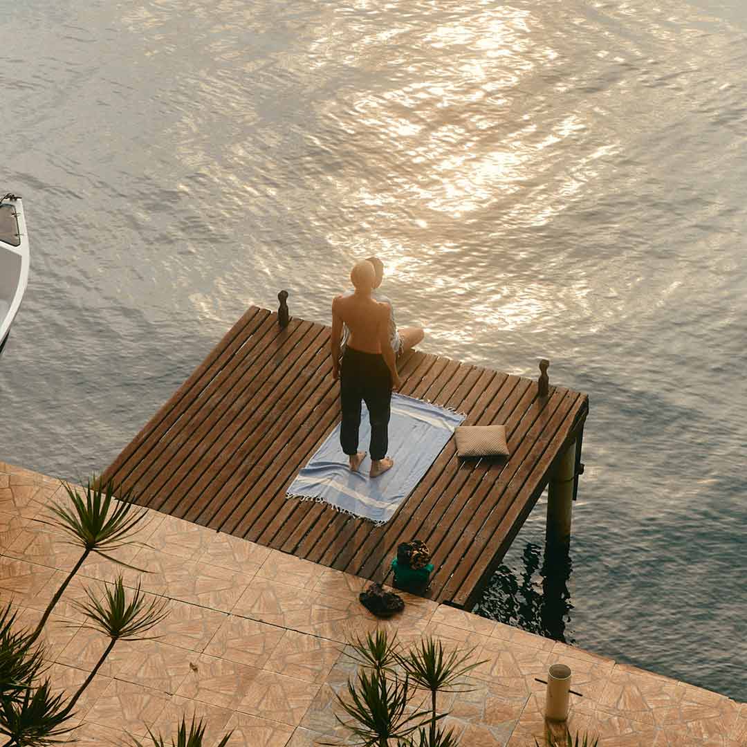 Eine Person steht entspannt auf einem hölzernen Steg und blickt über das ruhige Wasser im goldenen Abendlicht.