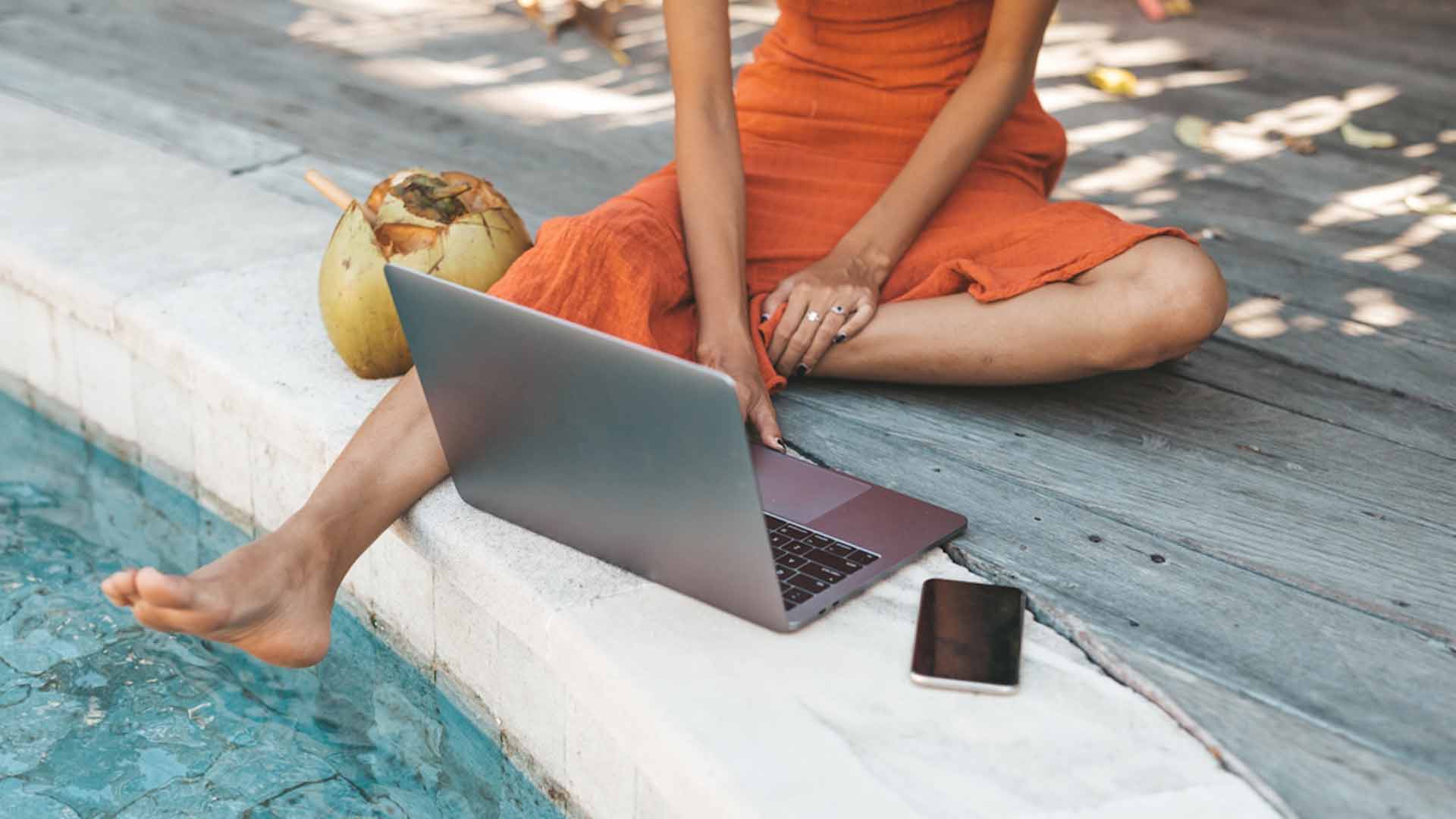 Person in an orange dress sitting by poolside with laptop, coconut drink, and smartphone nearby.