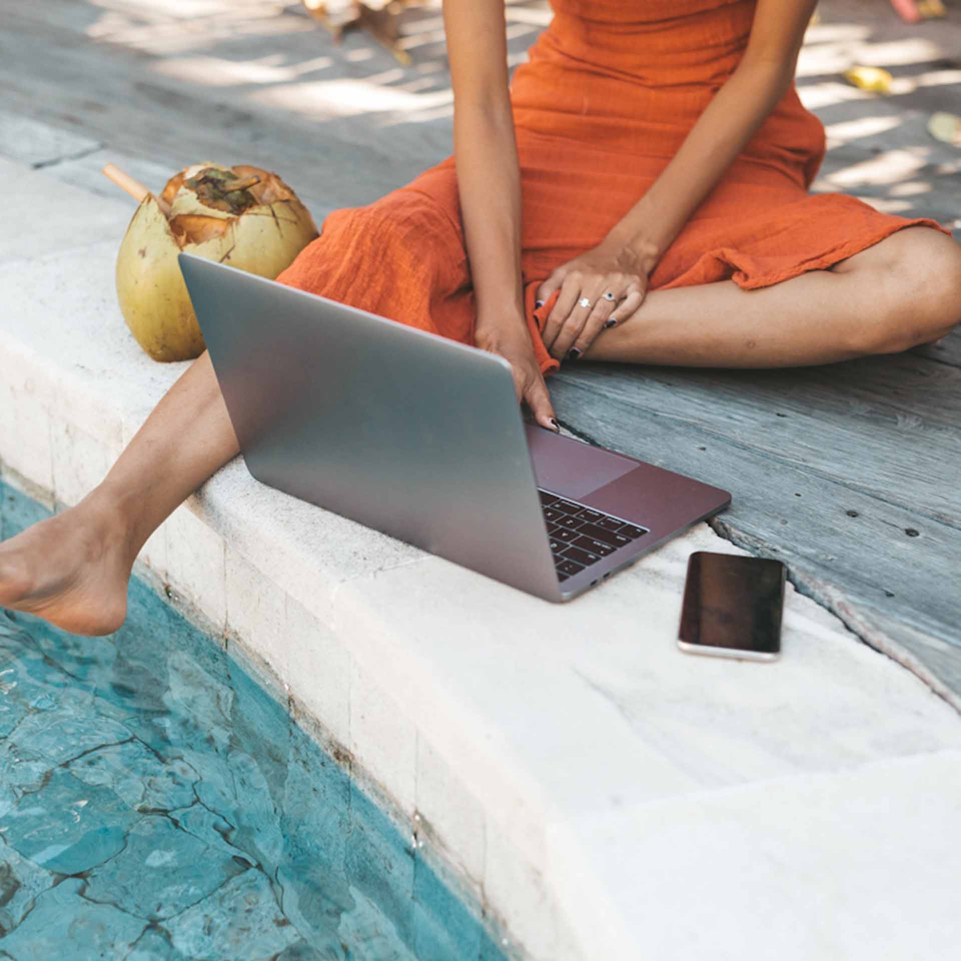 Woman in orange dress sitting by pool with laptop and smartphone nearby, barefoot with coconuts.