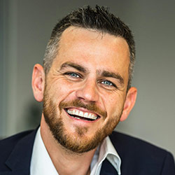 Smiling man with short dark hair, beard, and blue eyes wearing a dark jacket and white shirt.