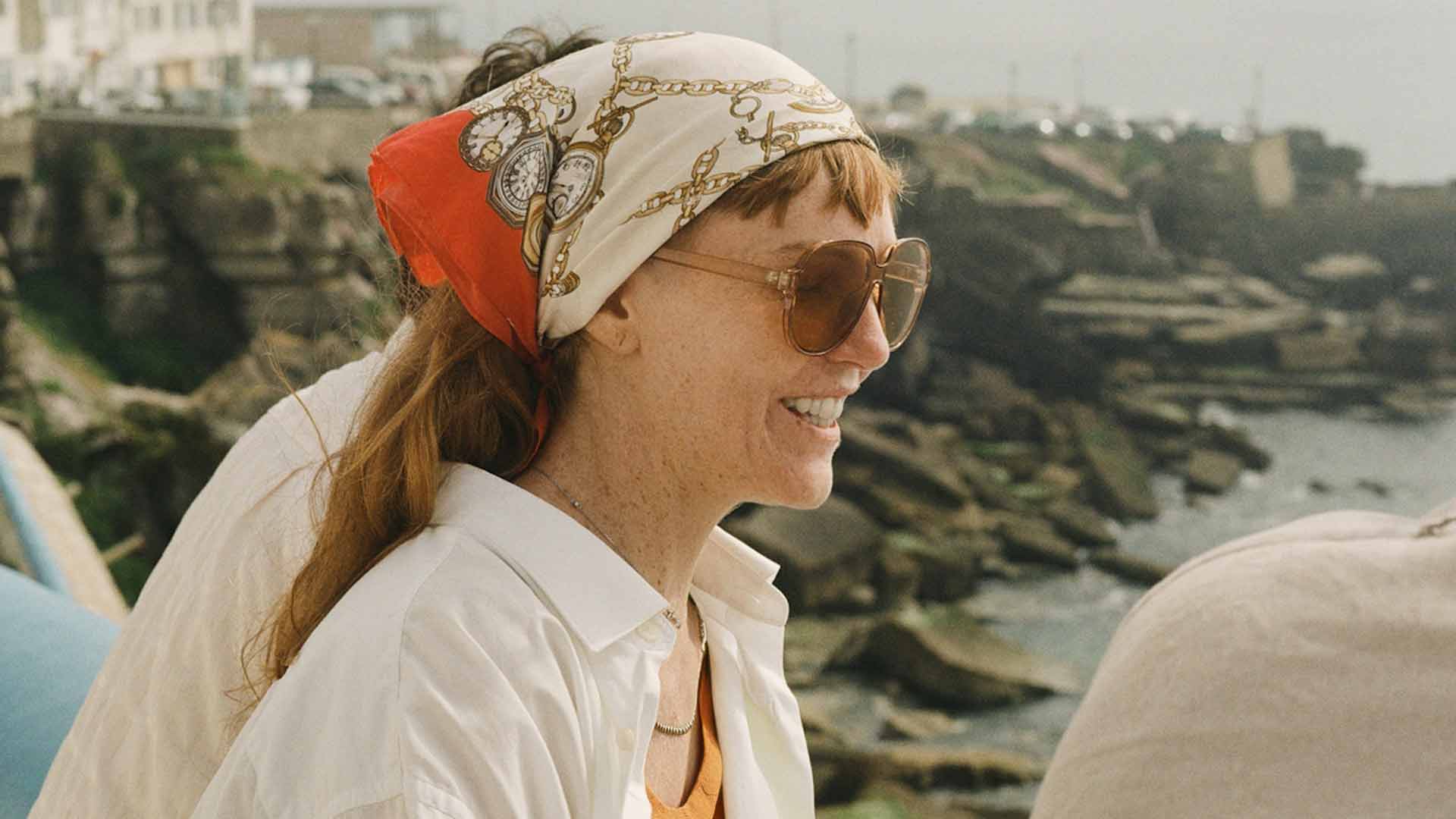 Smiling woman with headscarf and sunglasses near rocky seaside wearing white shirt and orange top.