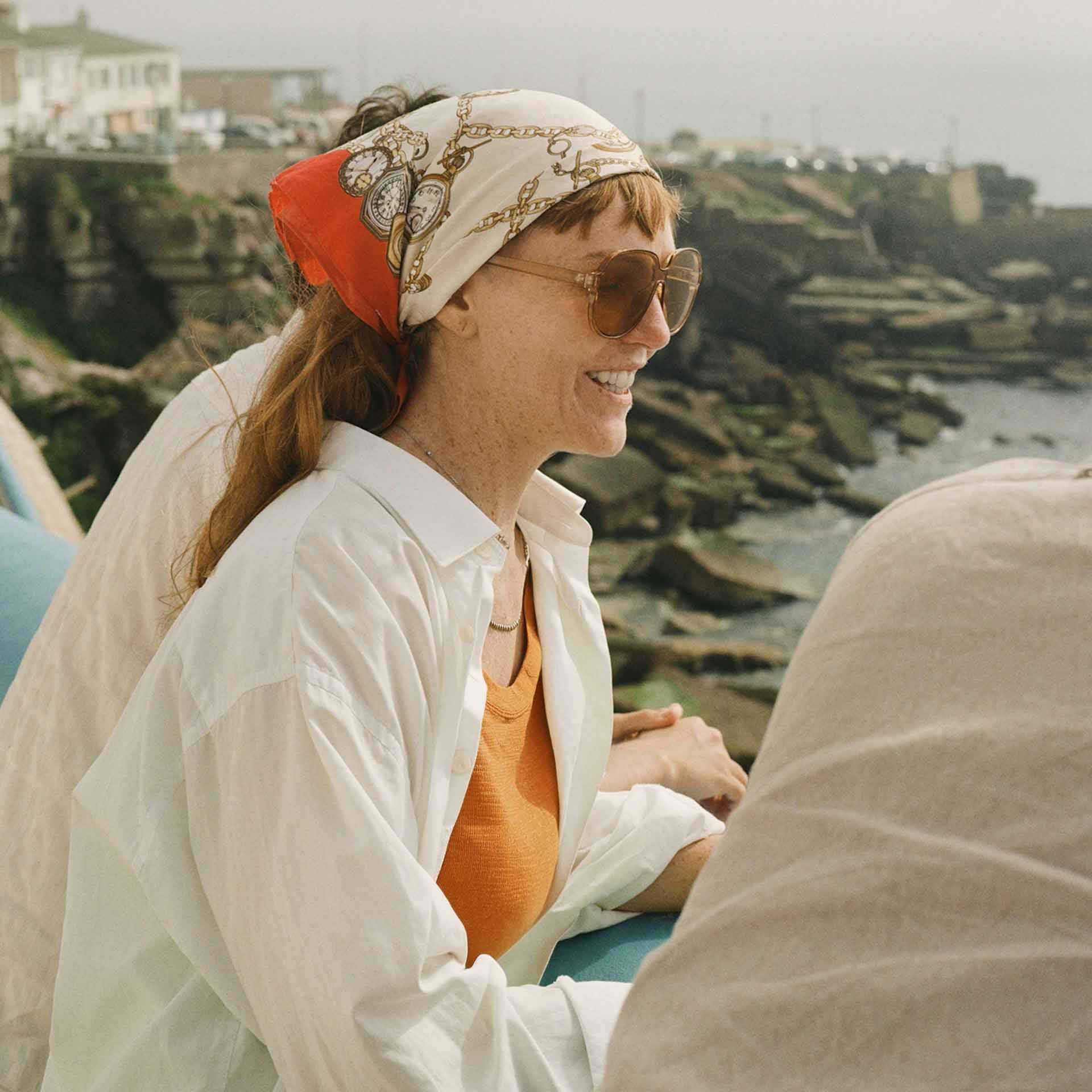 Smiling woman in sunglasses and a patterned headscarf leaning on a railing by rocky coastline.