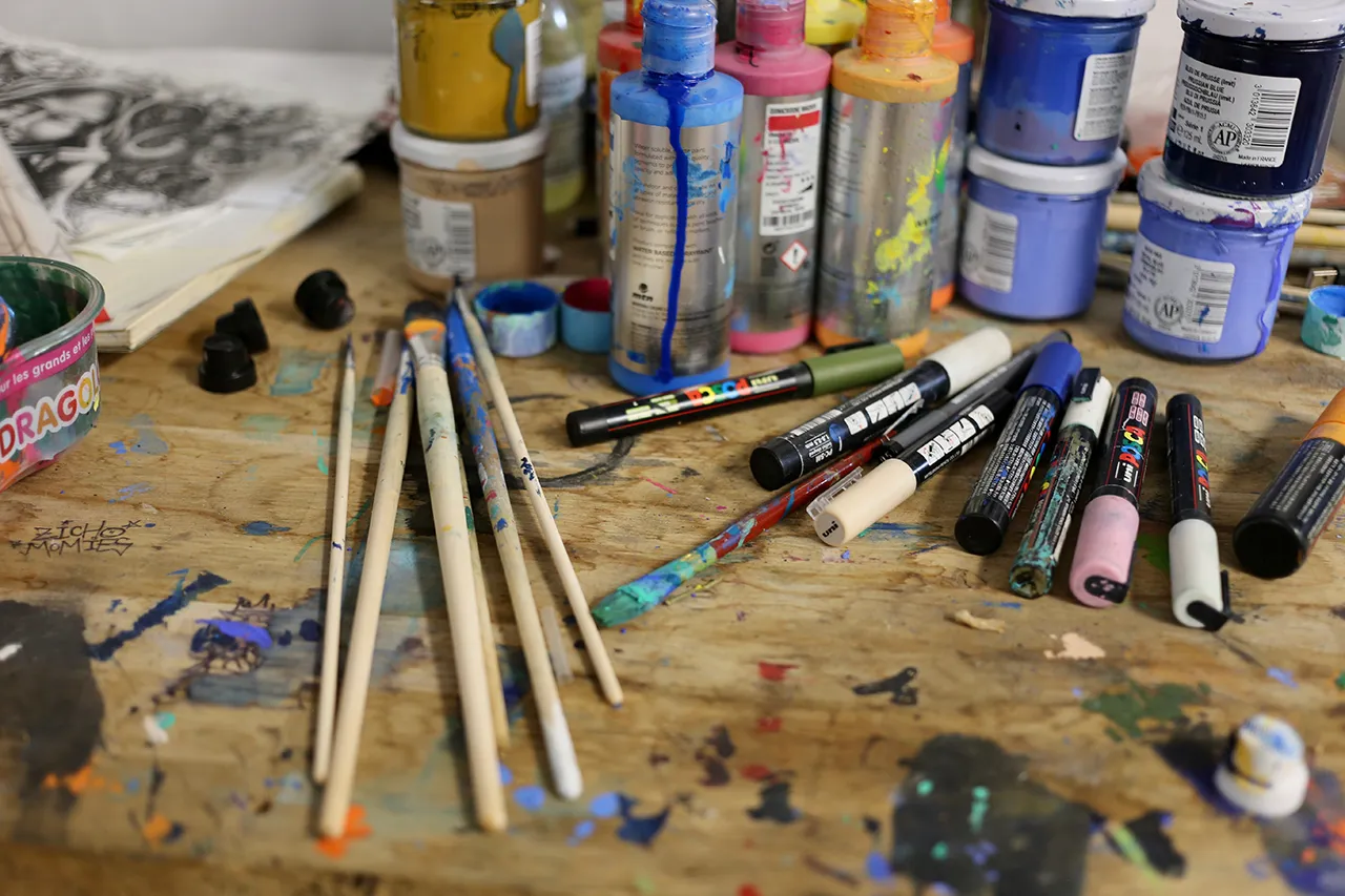 Plan rapproché d'un plan de travail d’atelier avec des pinceaux, des marqueurs et des pots de peinture aux couleurs vives, marqués de traces d'utilisation.