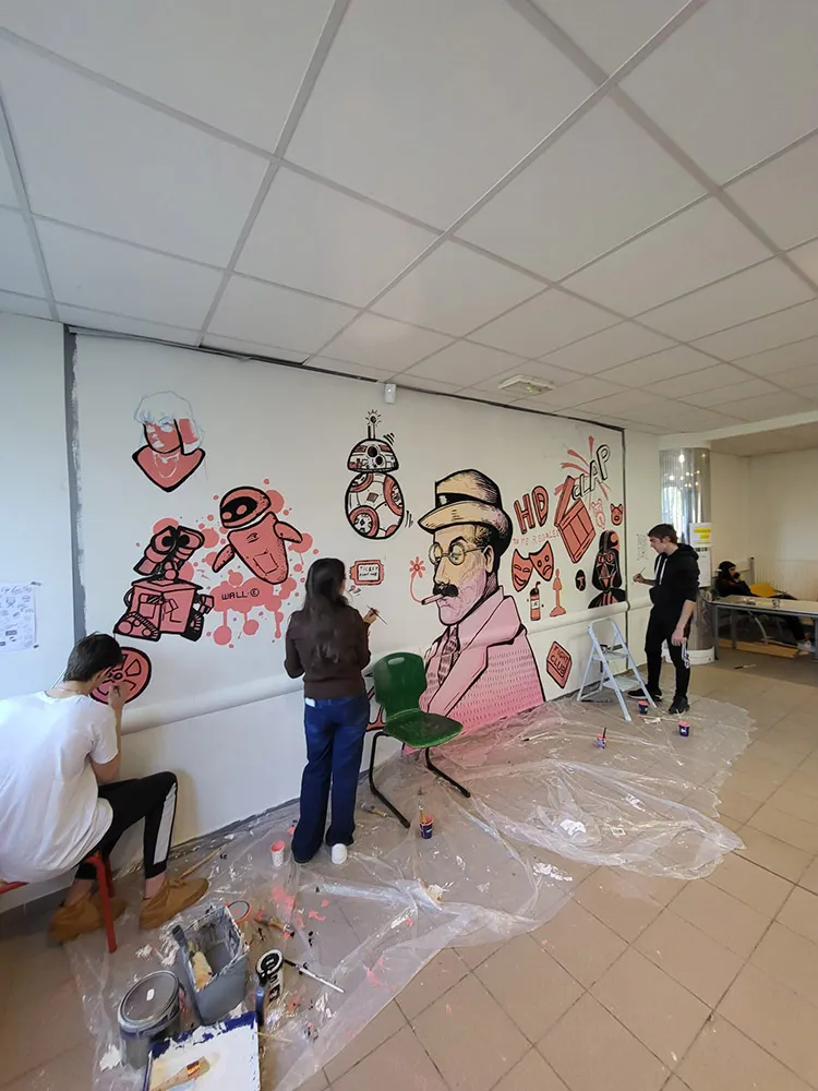 Plusieurs élèves assis et debout, peignant une fresque murale en rose et noir avec des motifs de cinéma. Des pots de peinture et du plastique protègent le sol.