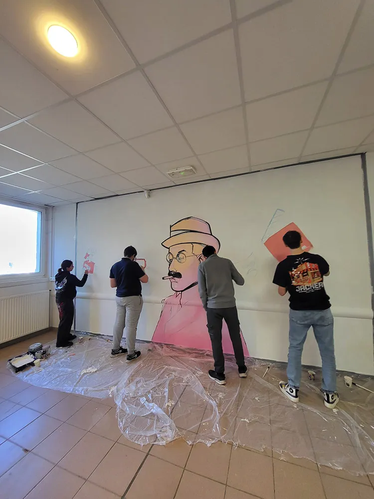 Plusieurs jeunes en train de peindre un grand mur blanc avec un portrait en cours de réalisation représentant un homme moustachu portant un chapeau, aux traits noirs et ombrages roses.