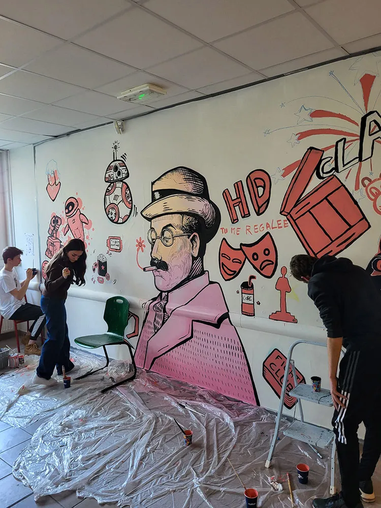 Un mur peint représentant un homme en rose et noir avec des lunettes et un chapeau, entouré de dessins inspirés du cinéma et de la pop culture. Une jeune fille et un garçon ajoutent des détails à l’œuvre.
