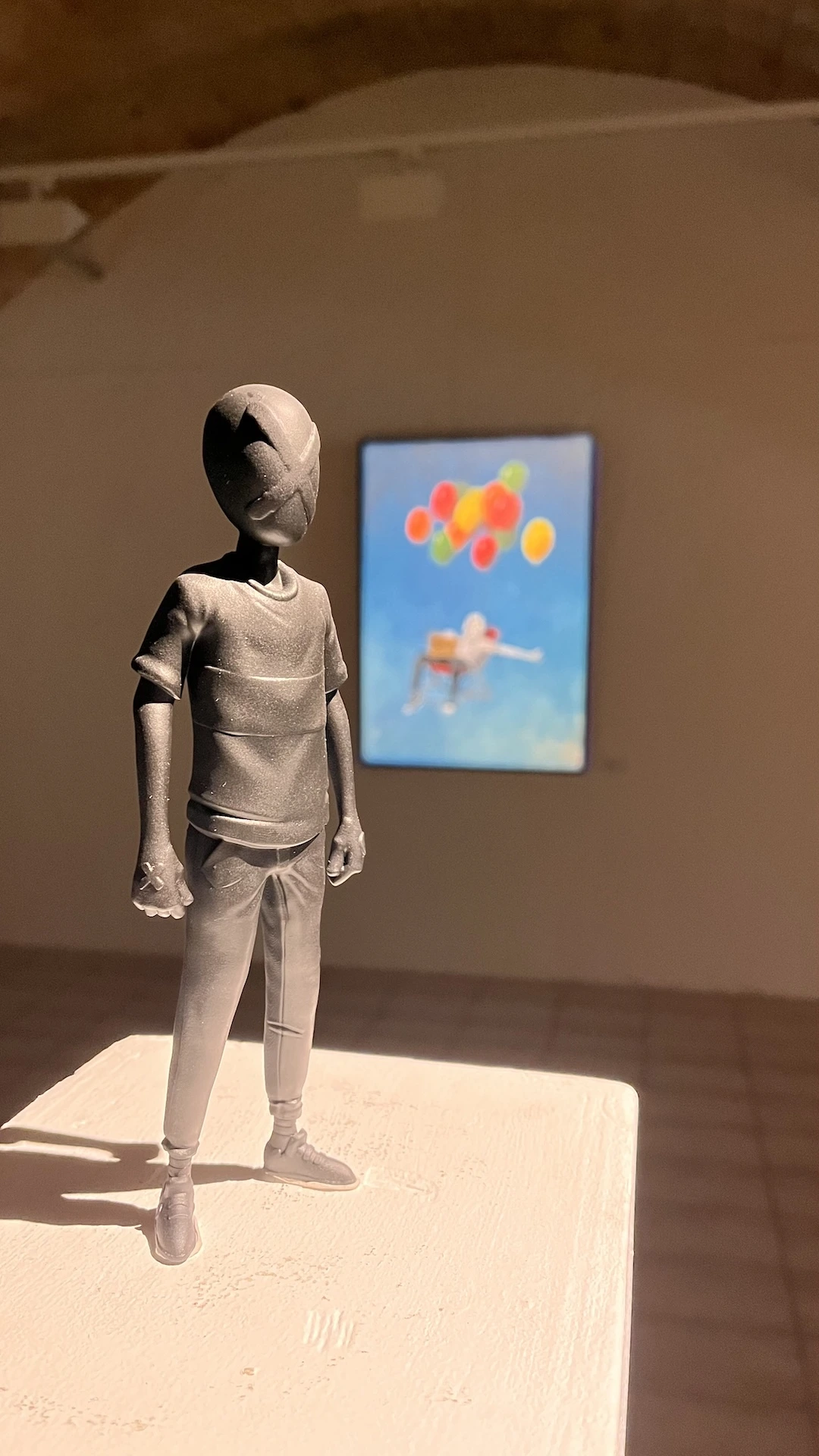 Figurine du personnage de Mara au premier plan, tout en noir, et au second plan une de ses oeuvres accrochée au mur.