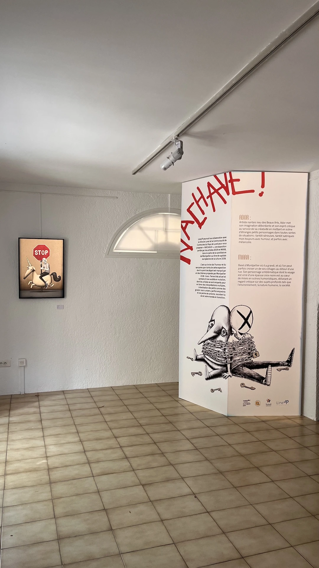Photo verticale montrant le paravent expliquant l'exposition, une oeuvre réalisée par les 2 artistes accrochée à côté.