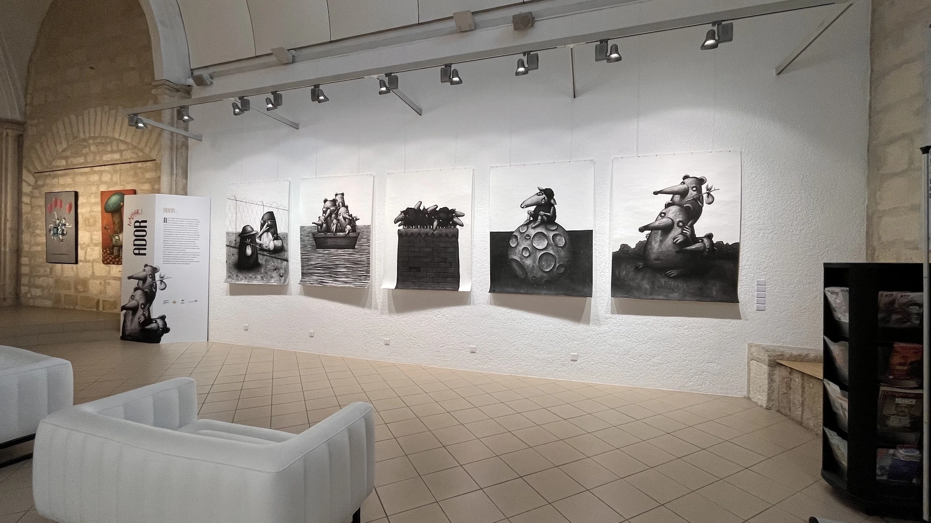 Suite de 5 grands dessins au fusain exposés sur un mur blanc, on voit des sièges blancs au centre de la pièce.