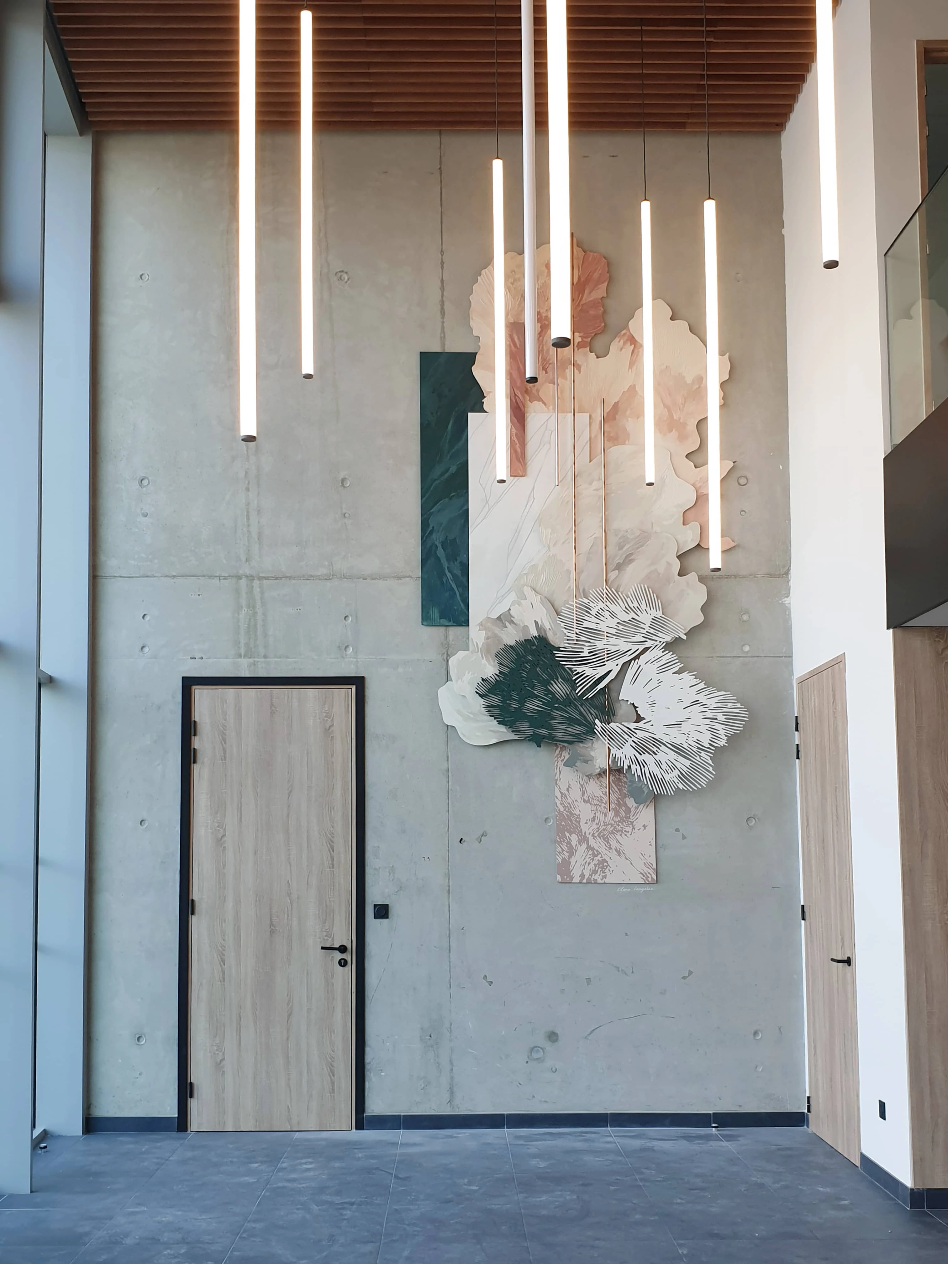 Photo d'un hall d'entrée avec du béton brut au sol et au mur, des luminaires verticaux qui pendant du plafond et une oeuvre accrochée au mur.