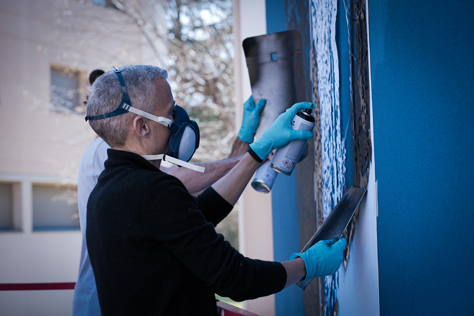 Photo de l'artiste portant un masque et des gants, une bombe de peinture à la main, entrain de peindre un mur.