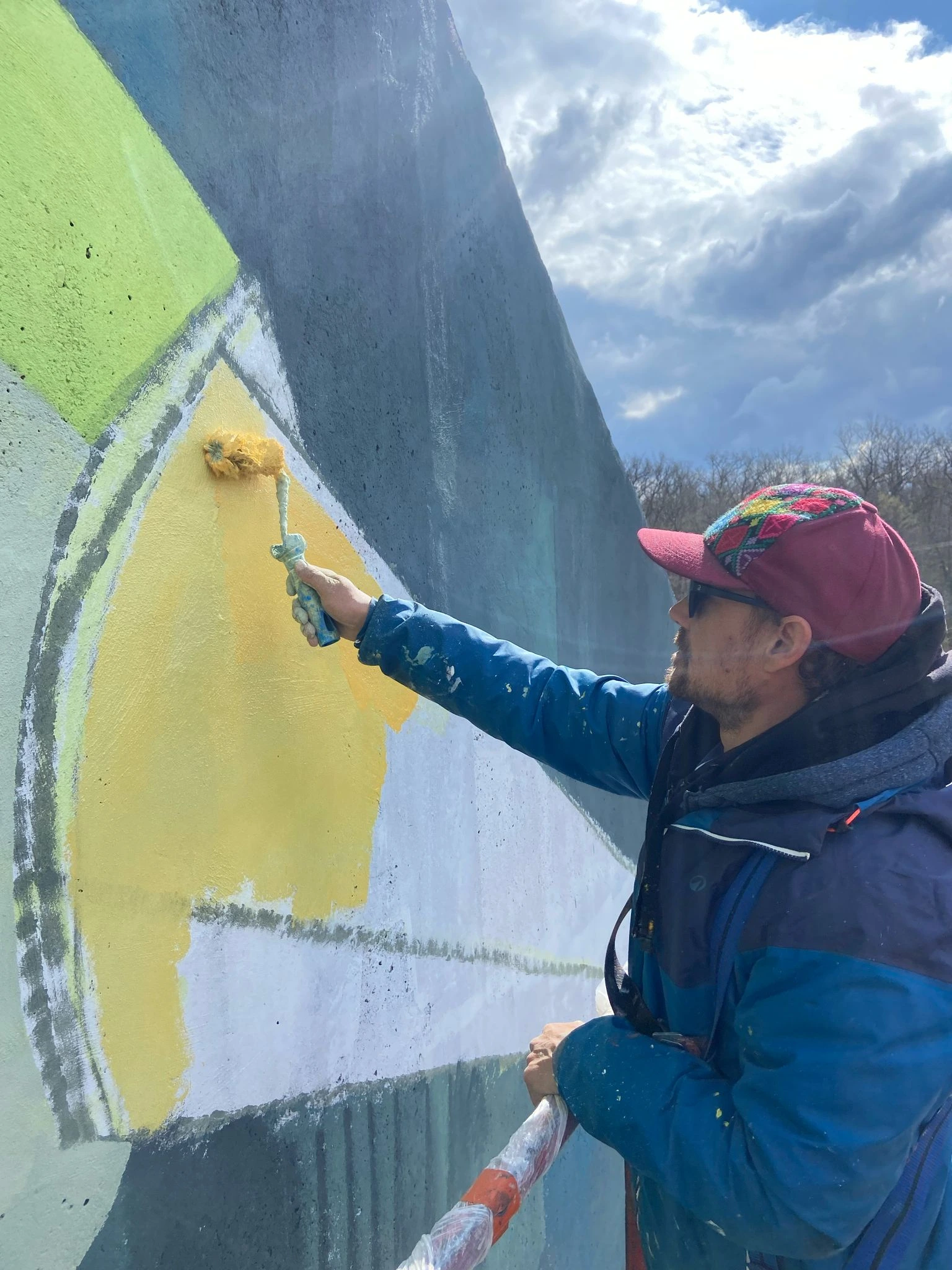Plan rapproché de l'artiste dans une nacelle entrain de peindre le mur, vêtu d'une casquette rouge et de lunettes de soleil.