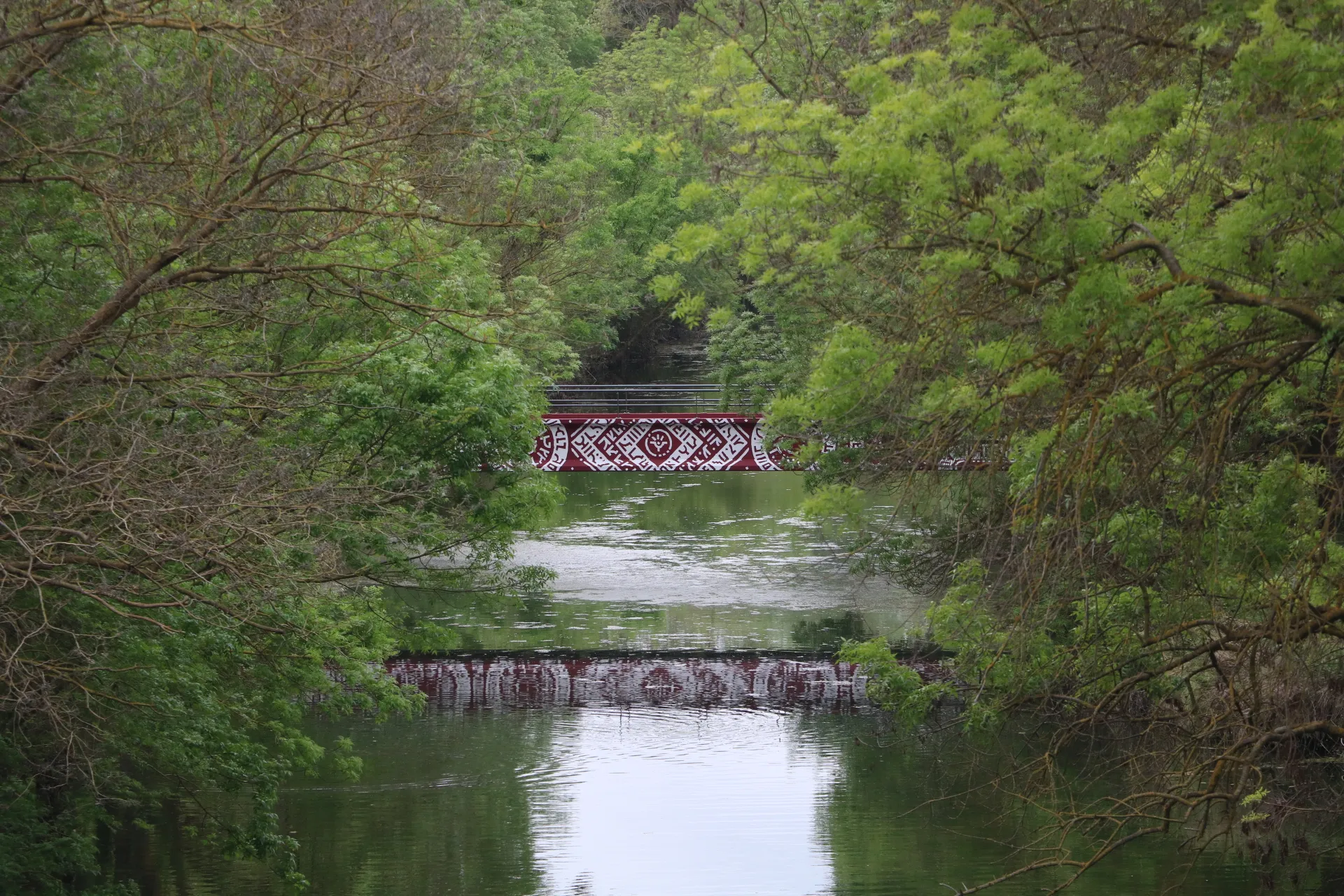 Vue lointaine d'une passerelle rouge décorée, entourée d’arbres et surplombant une rivière