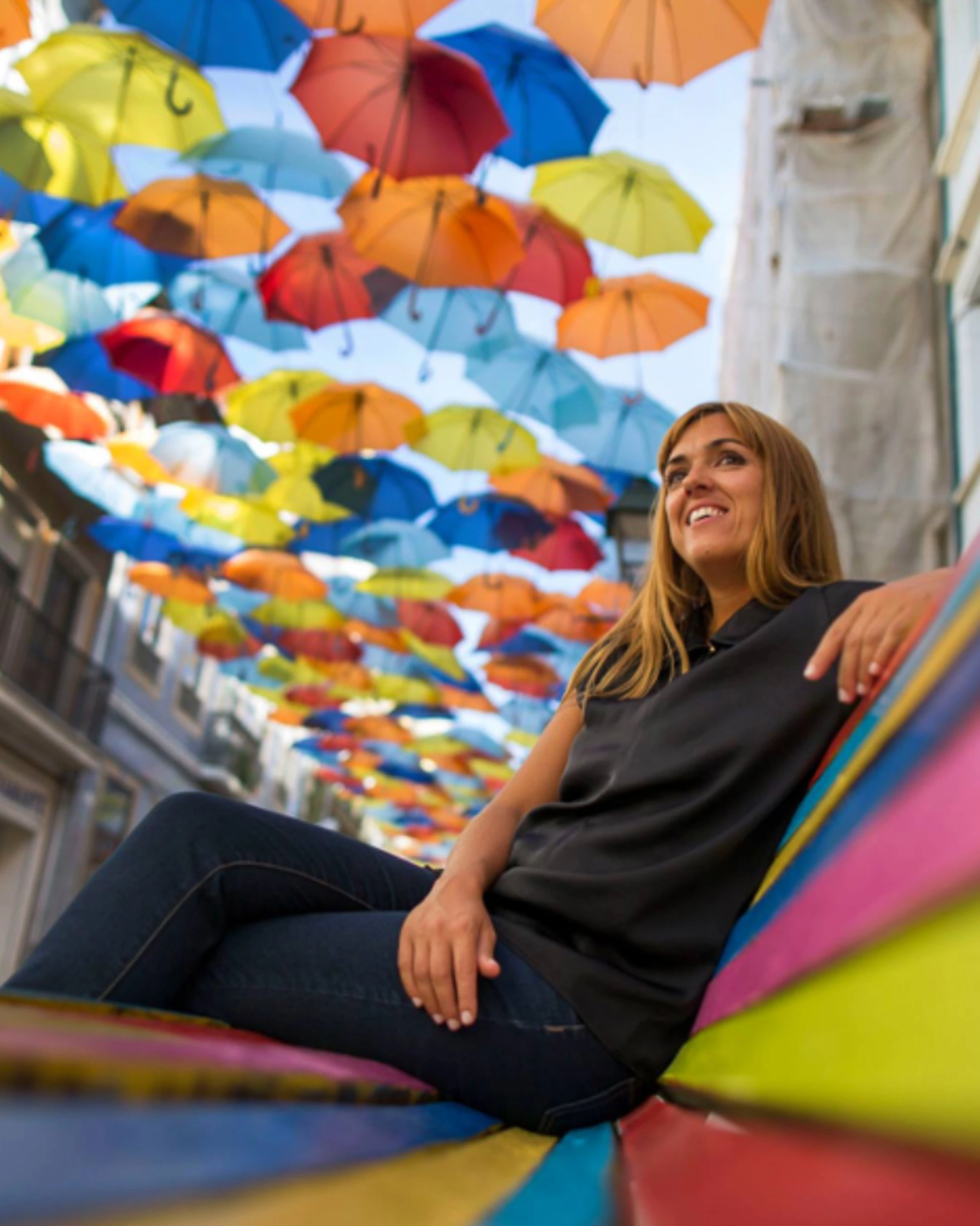 Photo de l'artiste assise sur un banc, en fond on voit des parapluies de toutes les couleurs suspendus dans les airs.