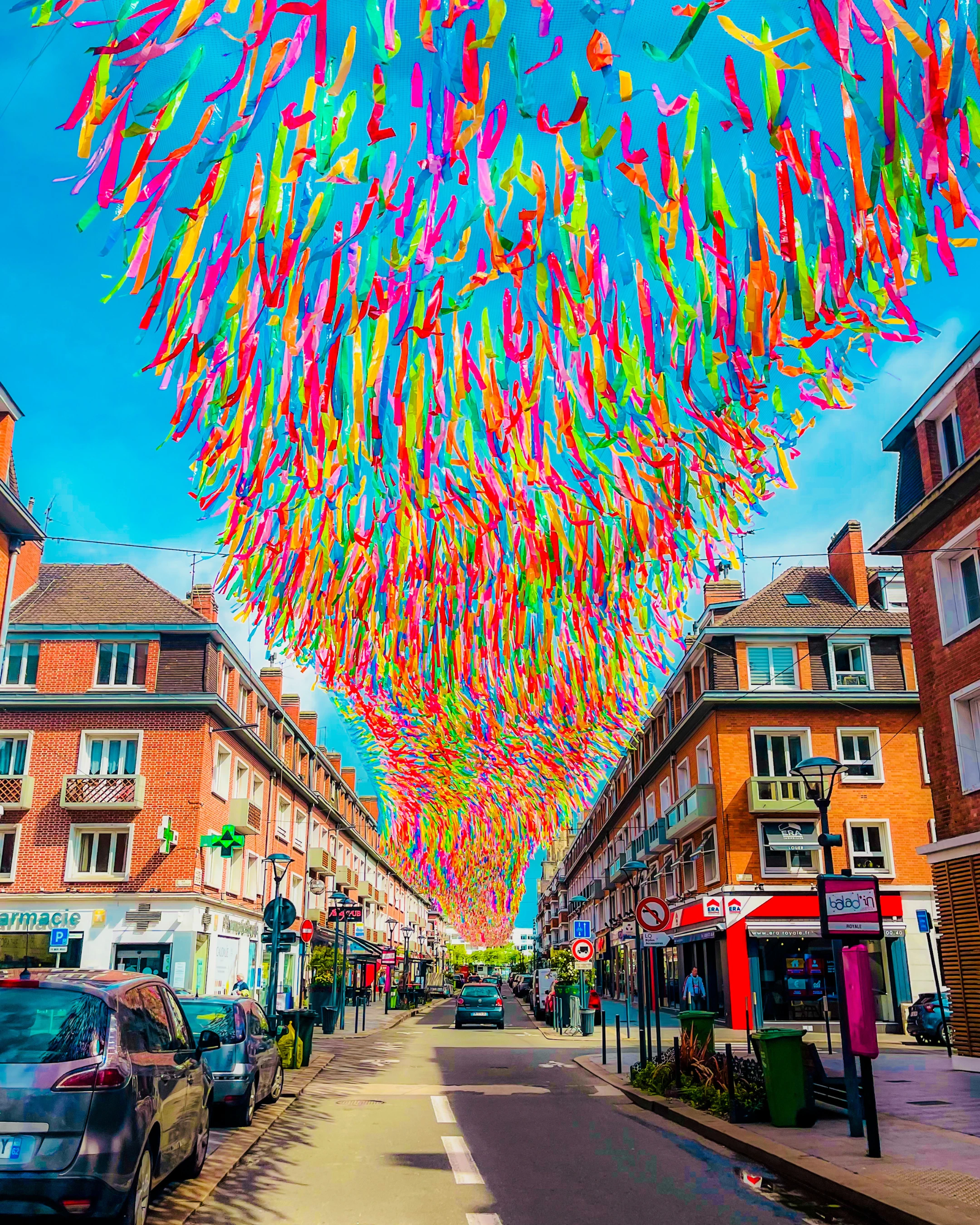 Installation artistique de rubans colorés suspendus au-dessus d'une rue bordée de bâtiments en briques rouges