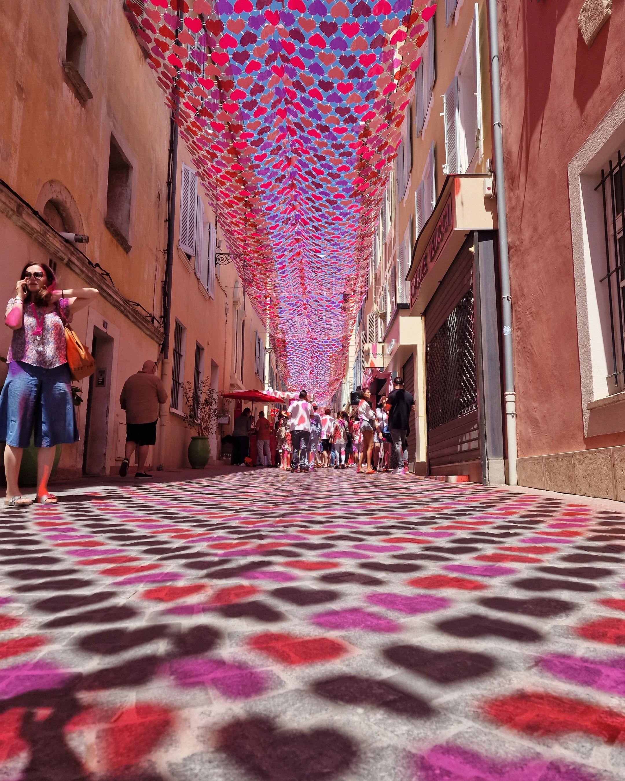 Rue étroite décorée de disques rouges et roses suspendus, créant des ombres colorées au sol