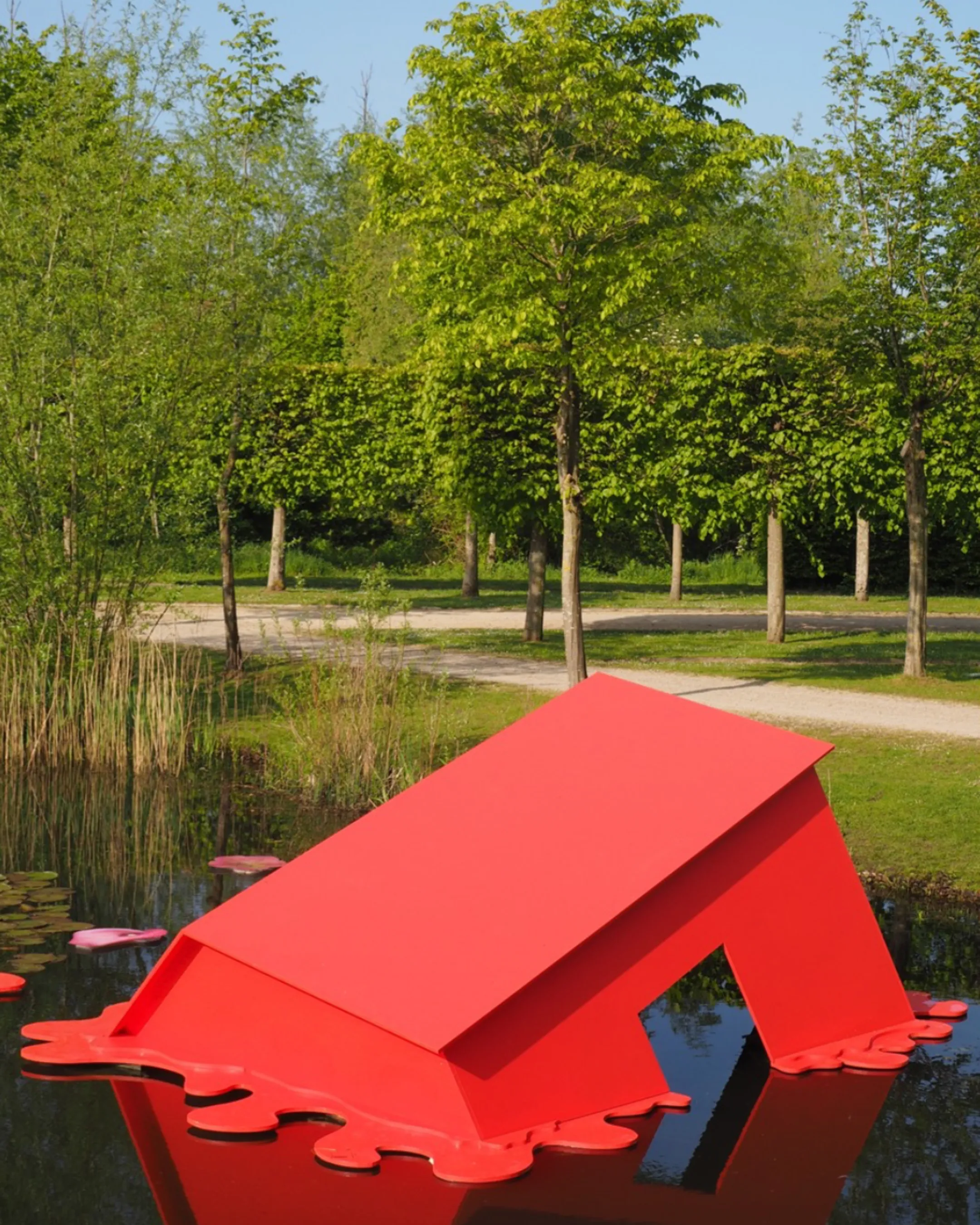 Sculpture contemporaine rouge posée sur l'eau, représentant un toit renversé, dans un parc verdoyant