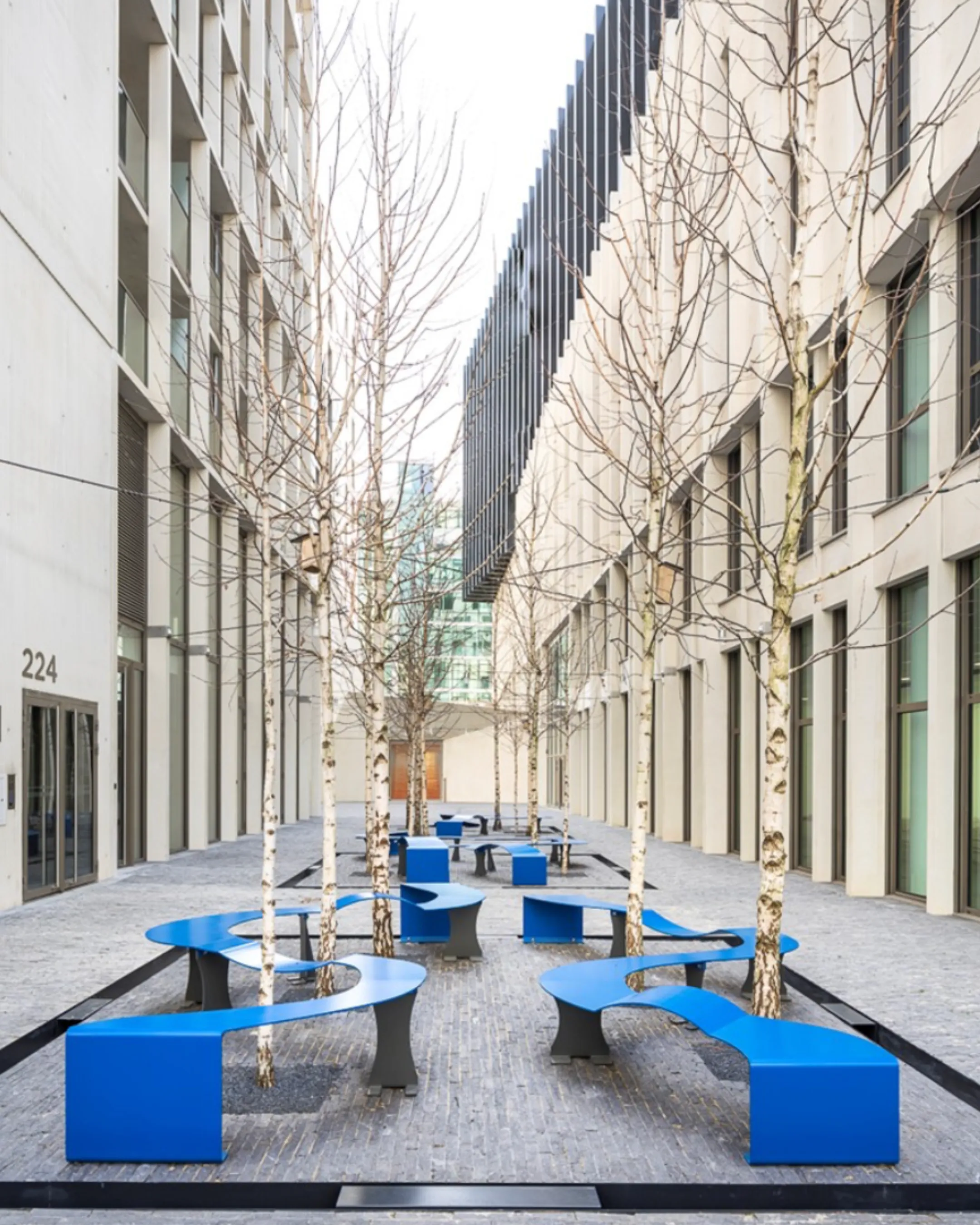 Mobilier urbain bleu disposé en formes géométriques dans une allée moderne bordée d'immeubles et de jeunes arbres