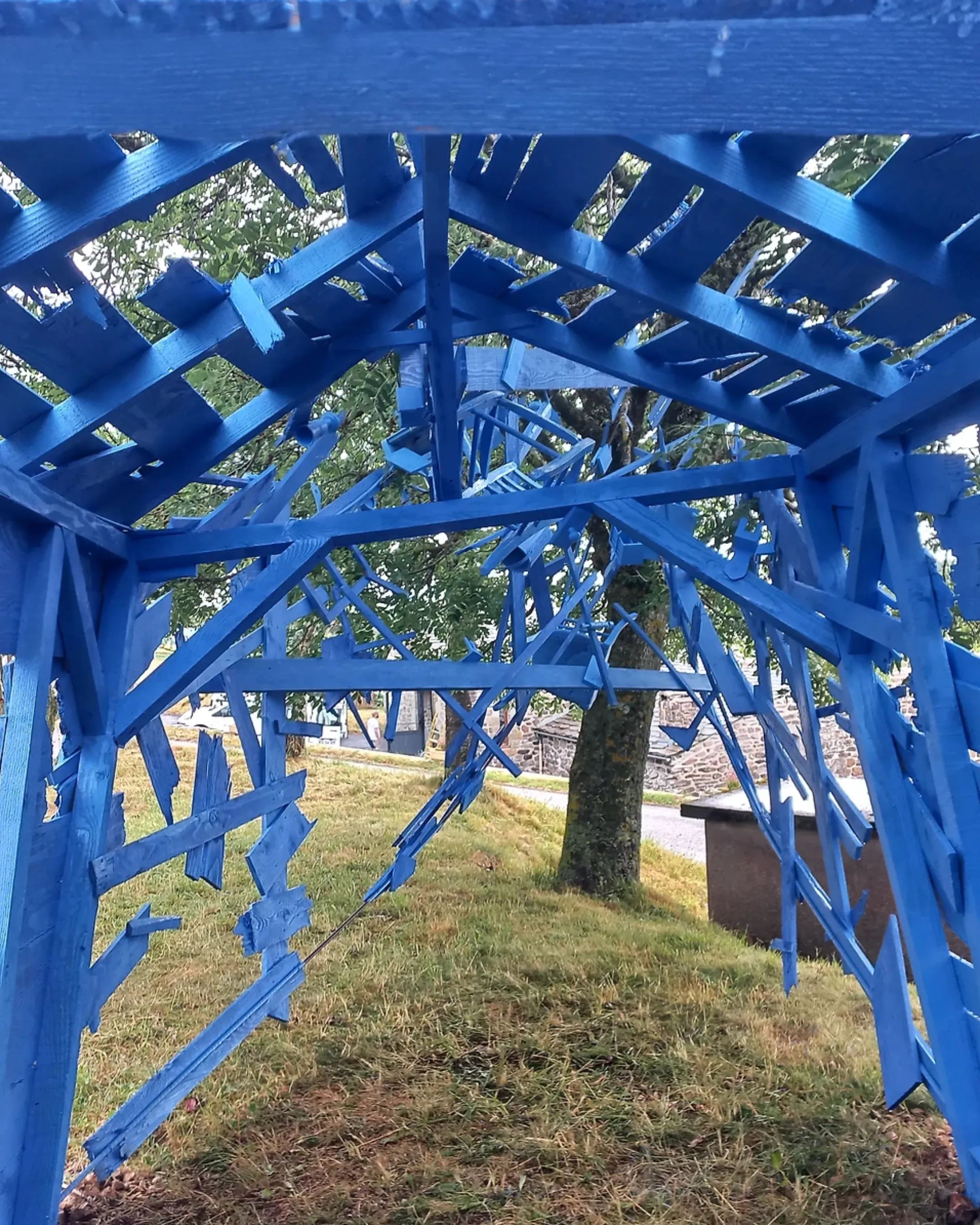 Structure en bois peinte en bleu, avec des découpes abstraites, installée sur un espace enherbé avec des arbres