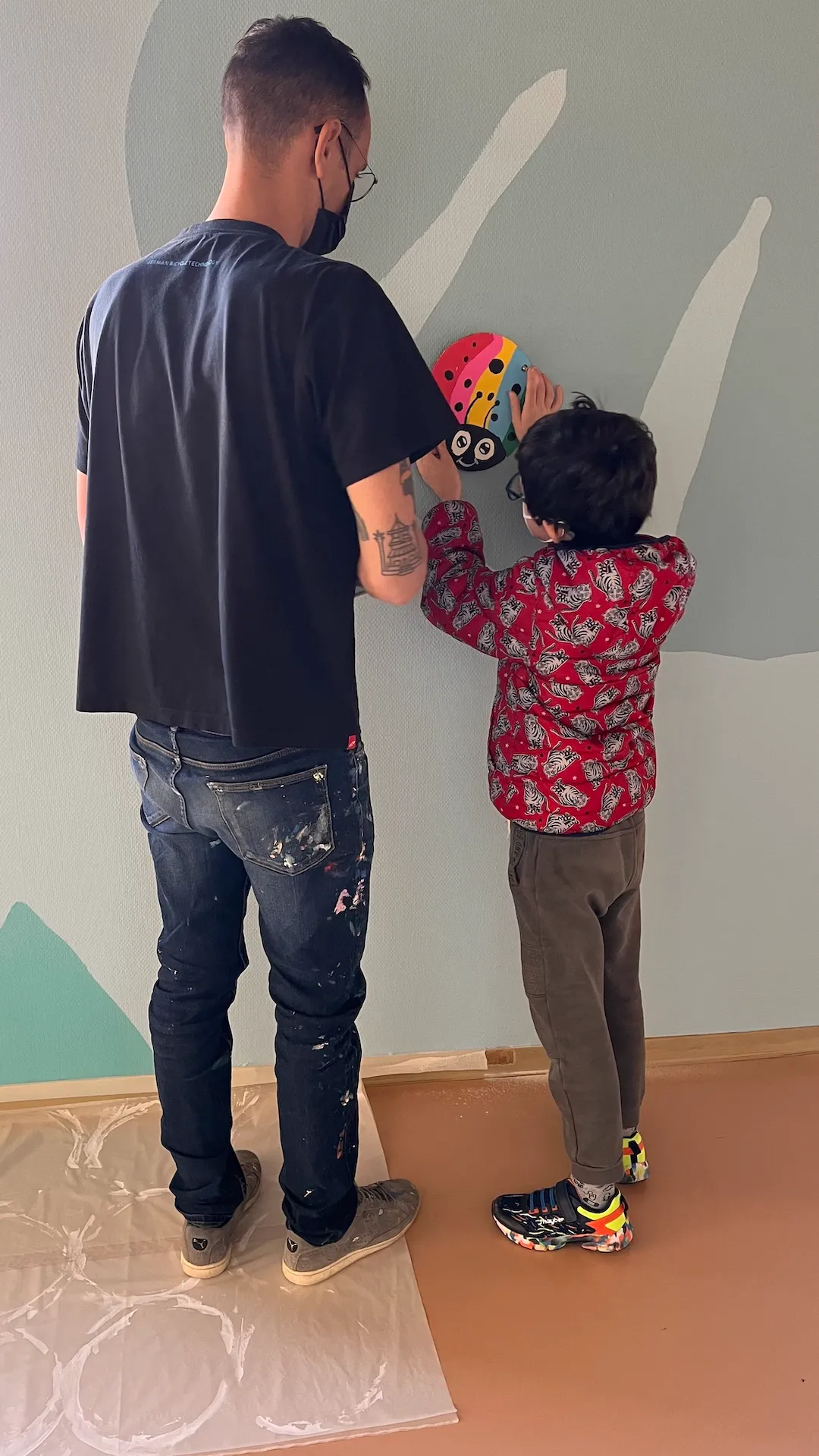 Photo de l'artiste entrain de placer un élément sur le mur, debout, avec un petit garçon qui l'aide.