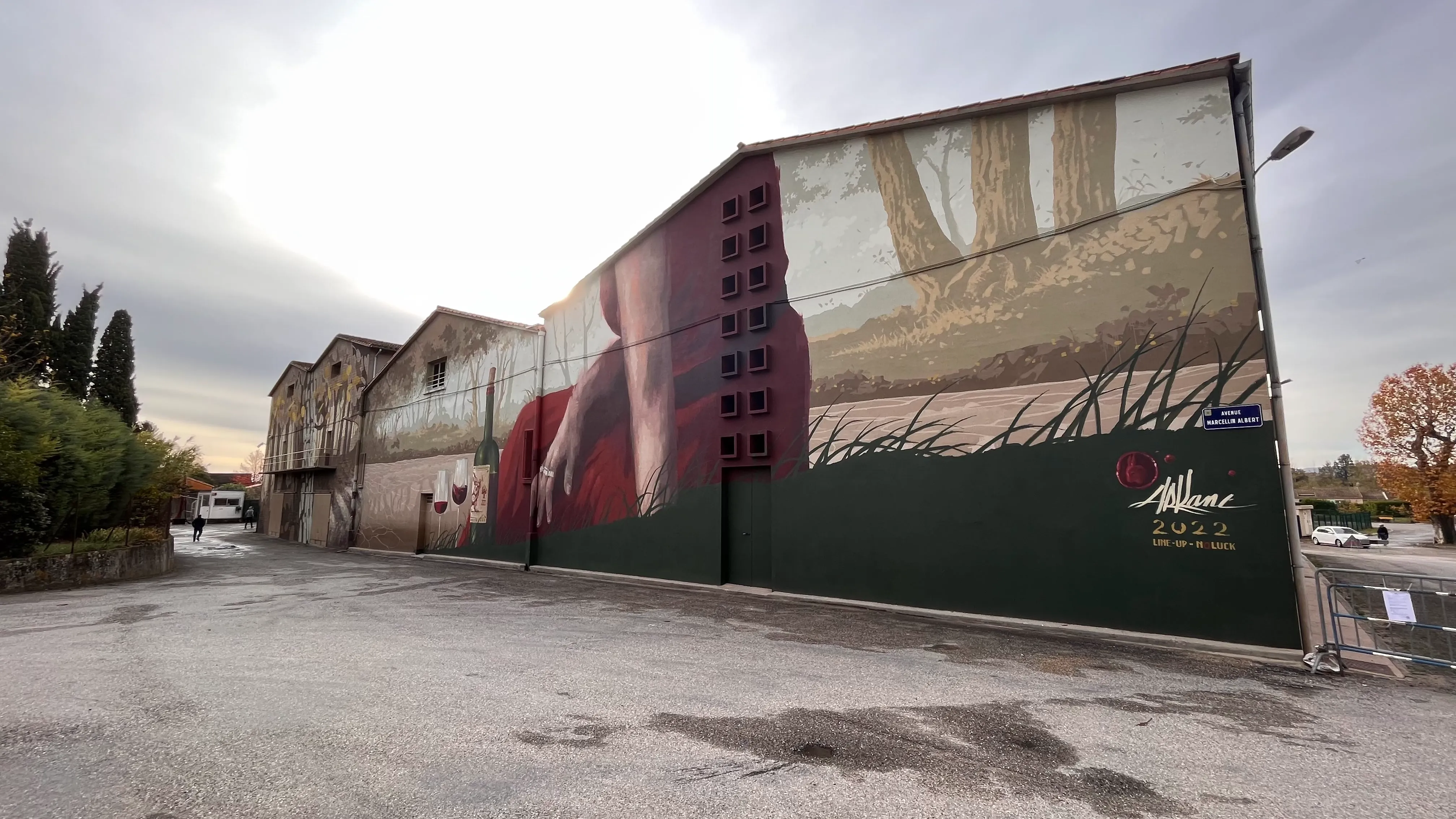 Vue large d'une fresque murale sur un bâtiment industriel, représentant des mains et du vin.