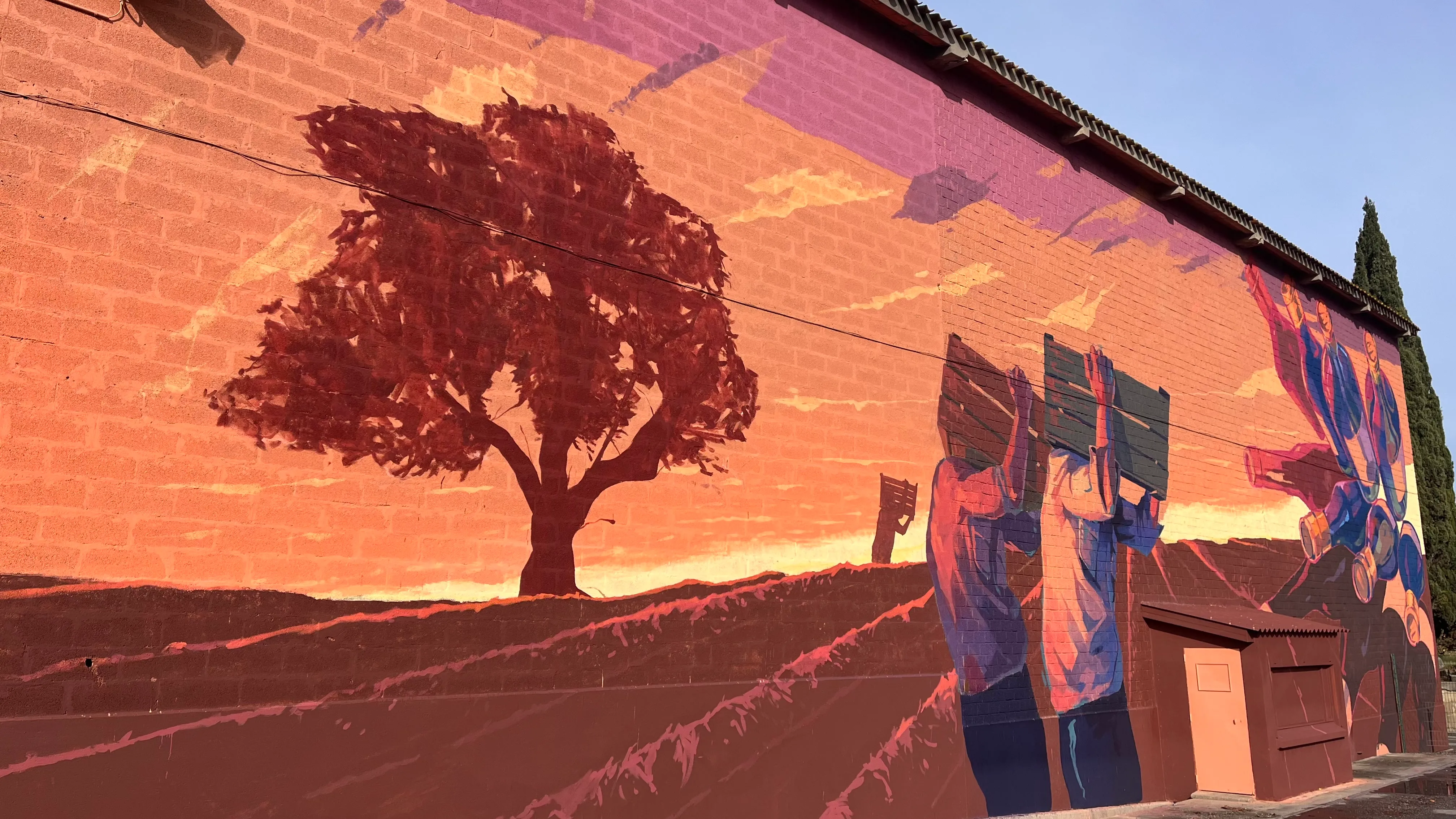 Détail de fresque murale montrant deux personnages stylisés portant des cagettes sous un arbre.