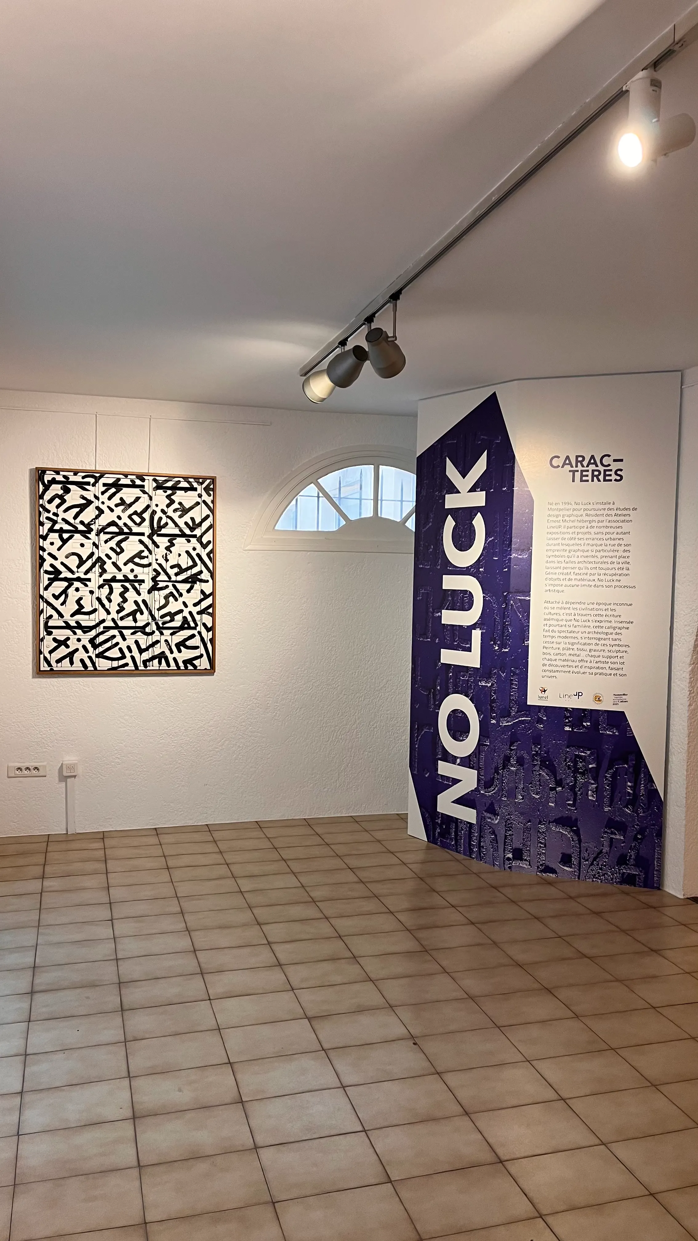 Œuvre en noir et blanc accrochée dans une pièce blanche, accompagnée d'un kakemono 'NO LUCK'.