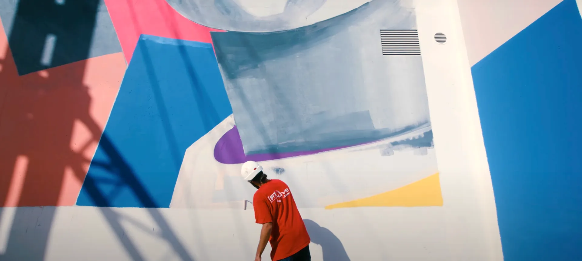 L'artiste, vêtu d'un t-shirt rouge et d'un casque de chantier blanc, et vu de dos entrain de peindre le mur.